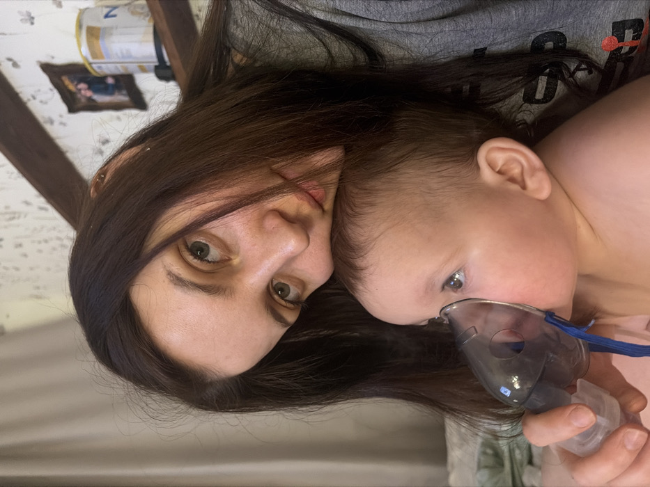 A woman with long, dark hair and large, green eyes holds a baby who is using a nebulizer. The woman looks directly at the camera with a concerned expression. The baby has fair skin and light-colored eyes, focused on something outside the frame. The background is slightly blurred, with hints of a room containing framed pictures and other household items. The photo appears to be taken indoors and at a close range.