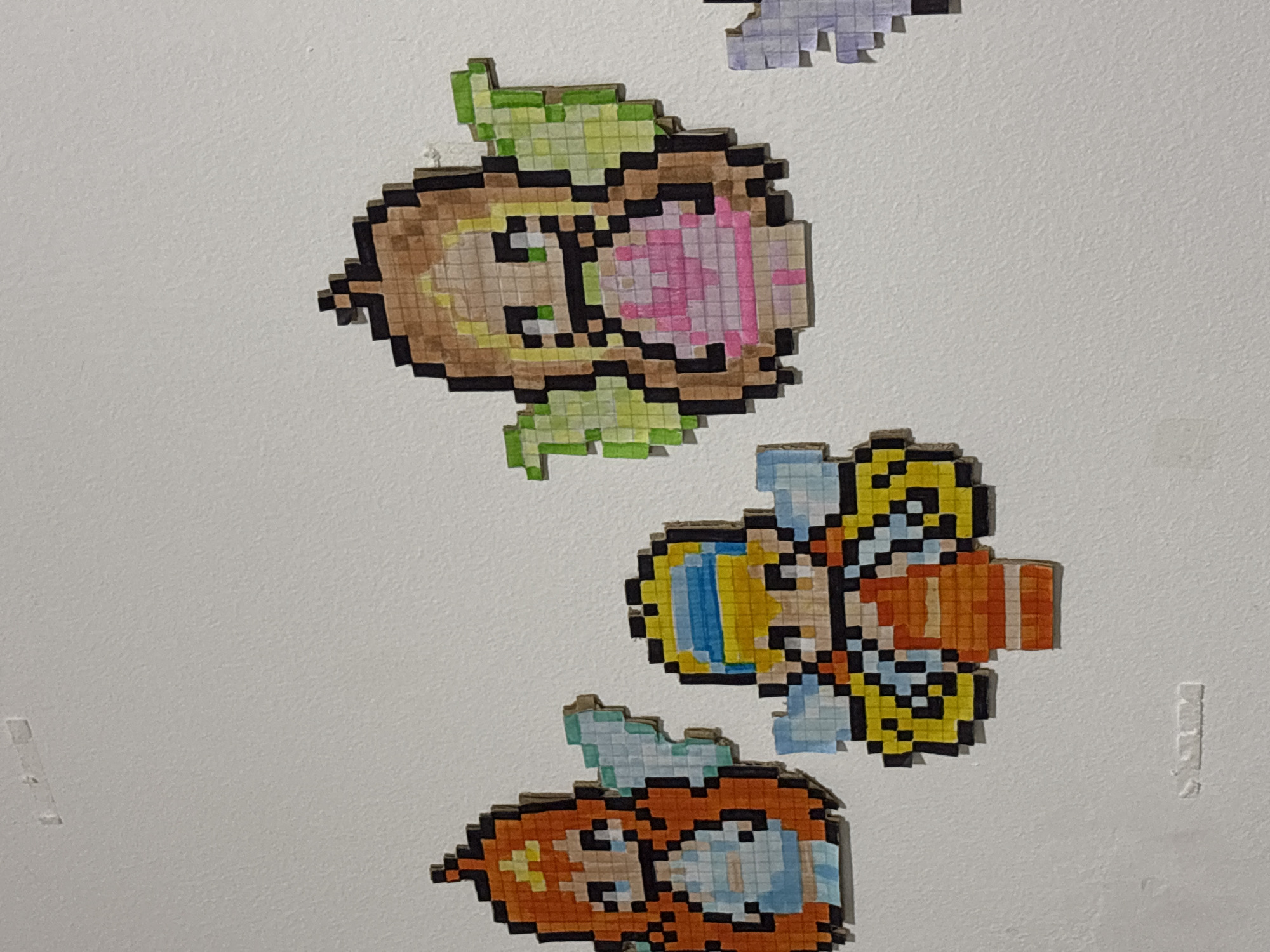 Three pixel art fairies are displayed on a white wall. The fairy on the left has orange hair and a blue dress. The fairy in the middle has blonde hair with blue accents in her hair and an orange dress. The fairy on the right has blonde hair and a pink dress. They are all attached to the wall with what appear to be small pieces of tape. There are also some illegible markings on the wall. The image does not contain enough information to determine its location in Fátima, Portugal.