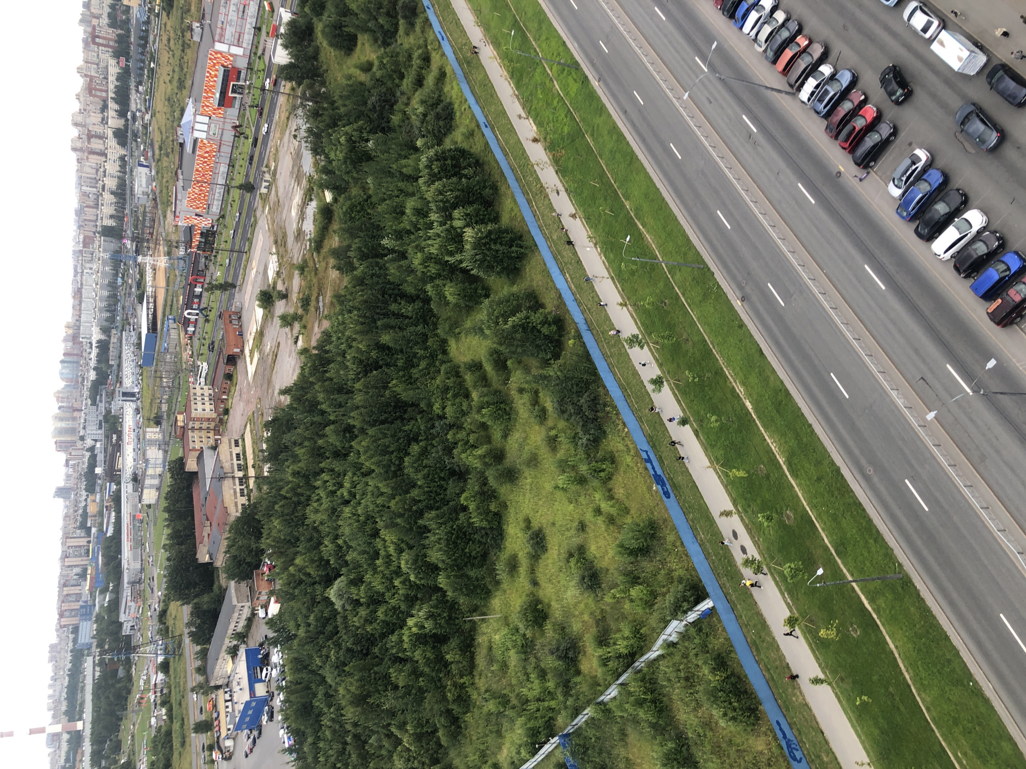 An aerial shot shows a wide, paved road with several lanes and parked cars. Adjacent to the road is a green grassy area, followed by a pedestrian walkway bordered by a blue fence. Beyond the walkway and fence is a dense forest, and in the distance, a cityscape with numerous buildings and industrial structures is visible. Several individuals are walking on the pedestrian path. The sky is overcast.