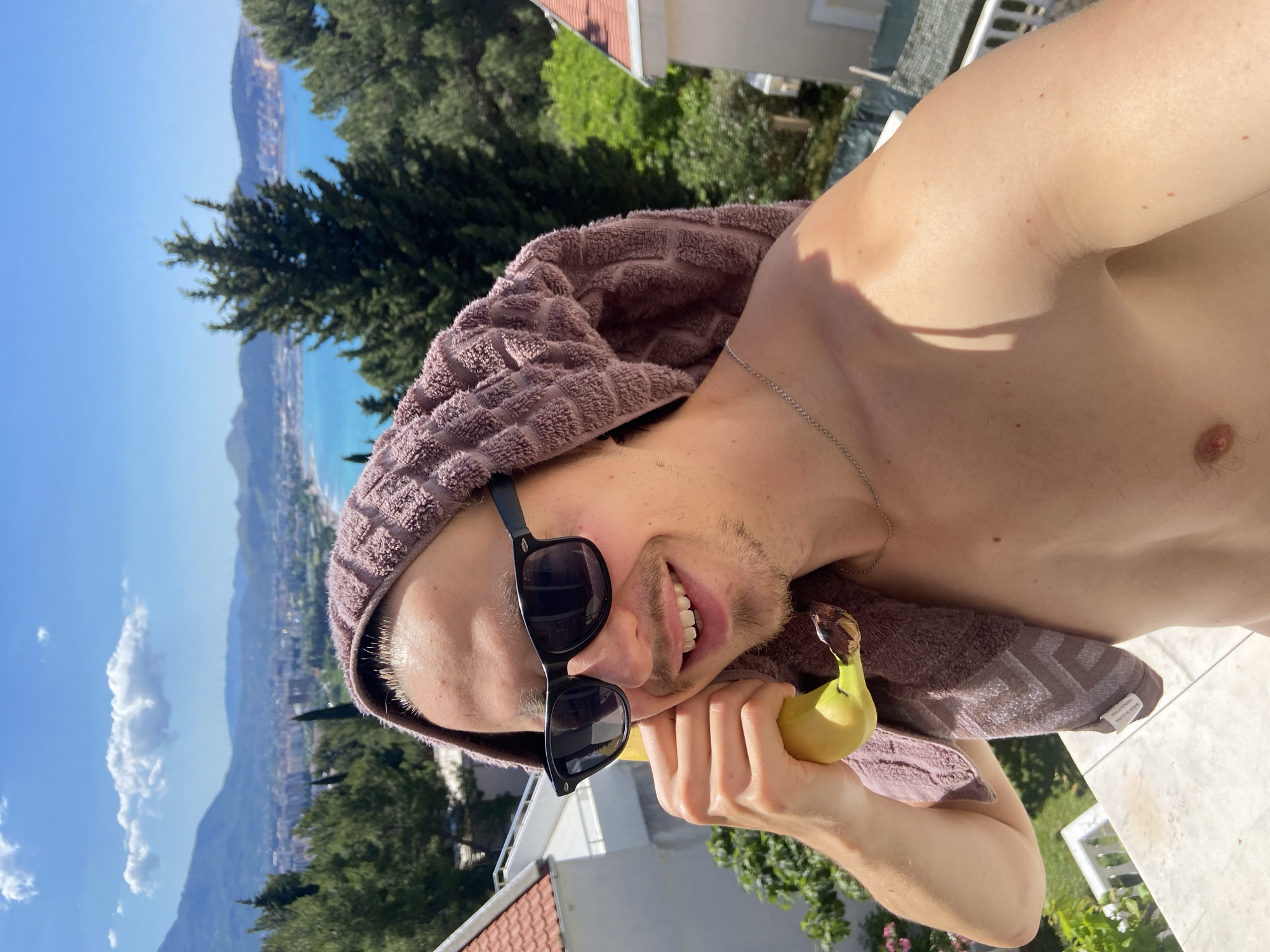 A man is holding a banana next to his ear, appearing to mimic using it as a phone. He is wearing sunglasses and has a brown towel draped over his head. He is shirtless and wearing a silver necklace. The background shows a view from a balcony or high vantage point overlooking the city of Šušanj, Montenegro. The scene includes buildings, trees, mountains, and a body of water under a clear, blue sky.