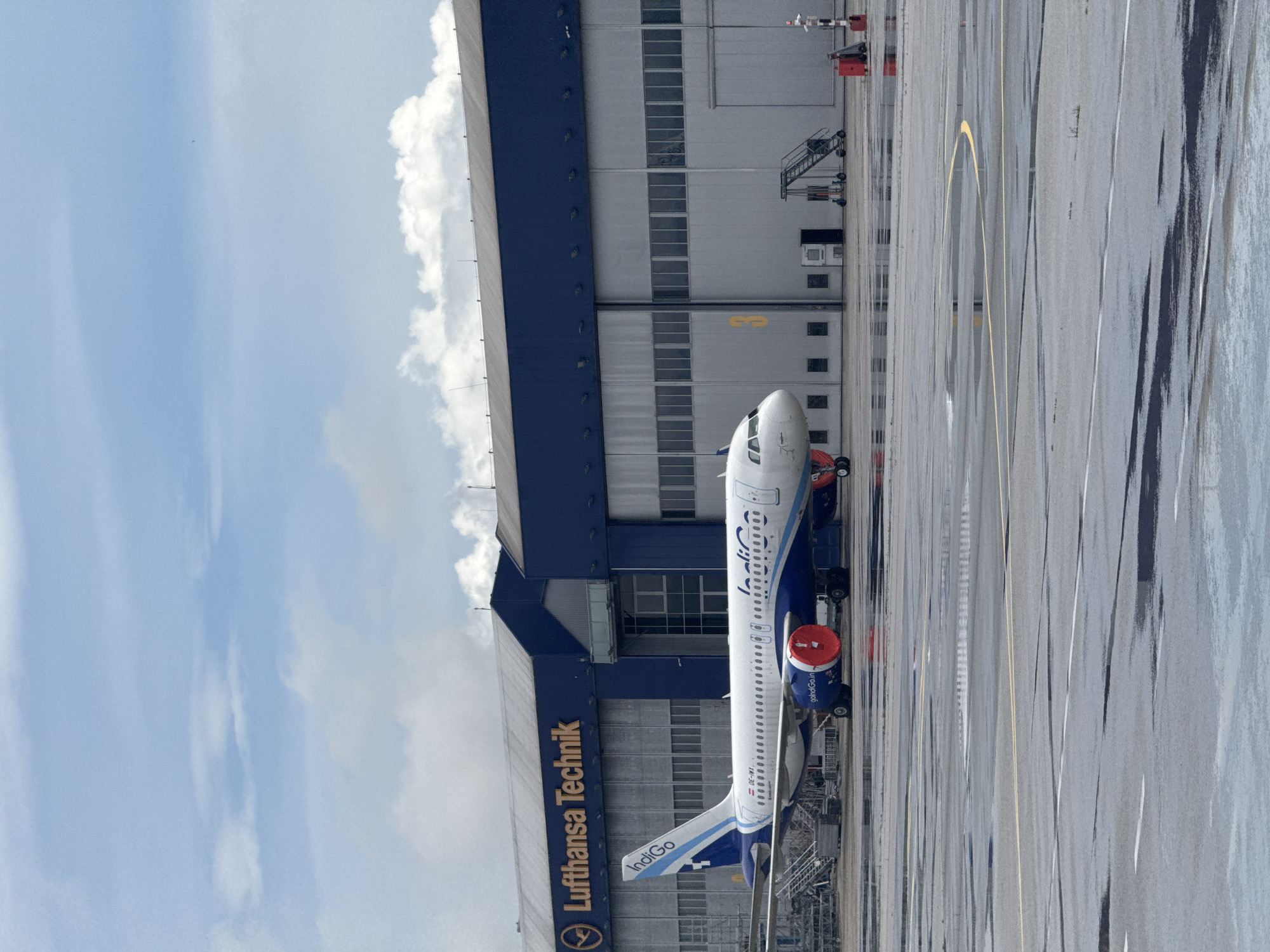 An IndiGo airline aircraft is parked on a wet tarmac next to a large maintenance hangar under a bright, partly cloudy sky. The image is taken during daylight hours.
The main subject is a white narrow-body passenger jet, an Airbus A320 family aircraft, featuring the blue and white livery of IndiGo. The airline's name, "IndiGo," is visible on the tail fin and along the fuselage. Red engine covers, with the text "goIndiGo" visible on them, are placed over both engines, indicating the aircraft is stationary, likely undergoing maintenance or being prepared for a flight. The aircraft's registration number, "OE-IMK," is visible on the fuselage near the tail.
To the left of the aircraft is a large, modern hangar building. The hangar has a distinctive dark blue upper section and a light grey, paneled lower section with multiple windows. Prominently displayed in gold lettering on the blue part of the hangar are the words "Lufthansa Technik," accompanied by the Lufthansa crane logo, indicating the facility is a maintenance base for the German airline's technical division.
The tarmac in the foreground and extending to the right appears wet, possibly from recent rain, reflecting the bright sky and the aircraft. Yellow and white ground markings for taxiways or parking positions are visible on the asphalt. The sky above is mostly clear blue with some scattered, fluffy white clouds. The overall scene depicts an operational airport environment, specifically a maintenance area, consistent with the location context of Sofia, Bulgaria.