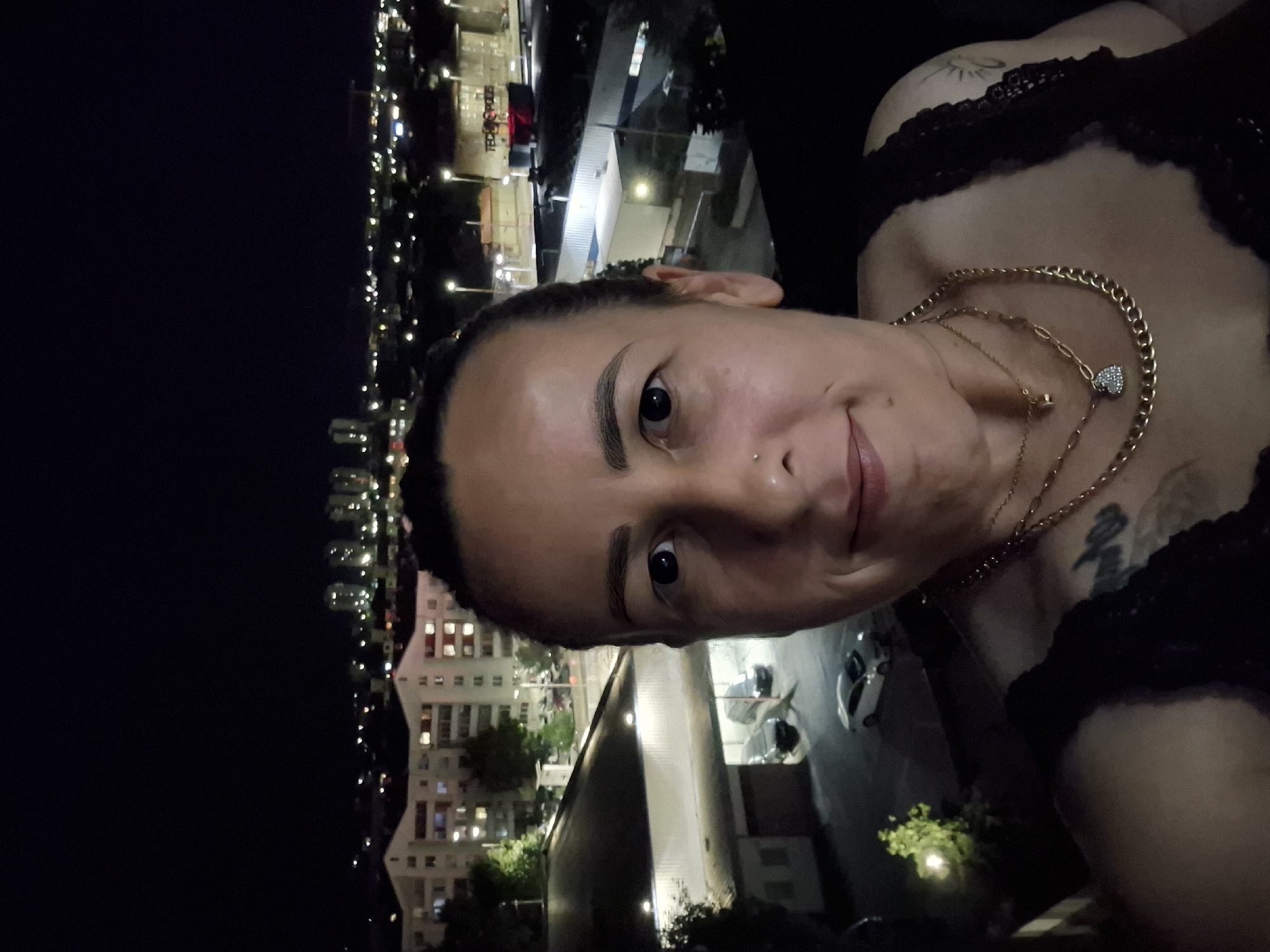 A woman is taking a selfie at night with an urban cityscape in the background. The woman is positioned in the foreground, with her face and upper body visible. She has dark hair pulled back, dark eyes, and is wearing multiple gold necklaces. She has a nose ring and visible tattoos on her shoulder and chest area. The background shows illuminated buildings and streetlights, suggesting a city environment. There are also cars visible in the distance. The overall lighting suggests it is nighttime.