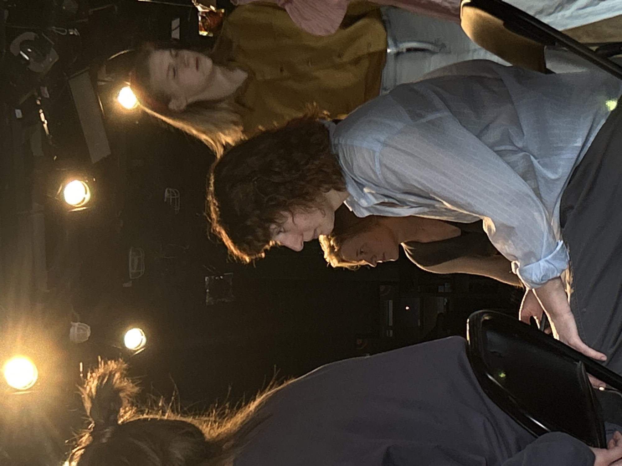 Four people are gathered indoors under stage lights. One person, with long hair, lies prone, wearing a mustard-yellow shirt. Another person is seated, leaning forward in a blue-striped shirt and grey pants. A third individual, wearing a black shirt, is angled towards the second person. The fourth person, wearing a dark-colored shirt, is bent over with their hair in a ponytail. The location appears to be a theatrical setting.