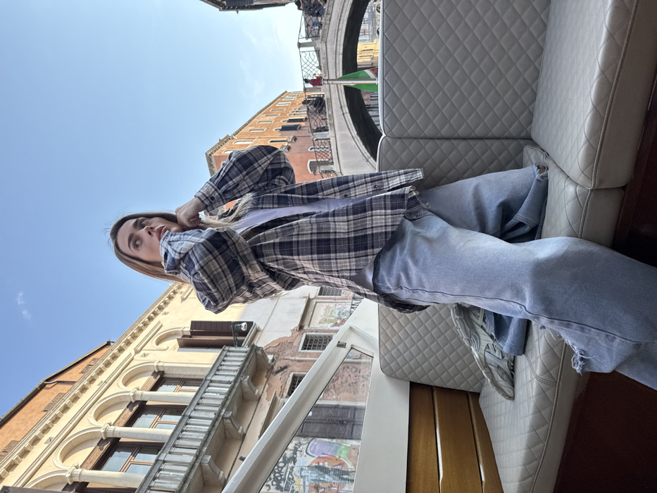 A young adult female with light brown hair is seated in a boat on a canal in Venice, Italy. She looks upwards and slightly to the right with an attentive expression, her hands raised to her upper chest, appearing to adjust her plaid shirt. She wears a blue, black, and white plaid long-sleeved shirt over a white t-shirt, paired with light-wash, wide-leg jeans featuring distressed details. She occupies a light grey, diamond-quilted upholstered seat within the boat, which also shows wooden paneling and a white frame of a canopy.
In the background, traditional Venetian buildings line a narrow canal under a clear blue sky. The architecture features multi-story structures with arched windows and balconies, displaying facades in tones of cream, ochre, and terracotta. One building shows signs of wear and graffiti at its lower levels. A stone arch bridge spans the canal in the middle distance, with several pedestrians visible on its surface. An Italian flag, green, white, and red, hangs vertically near the bridge.