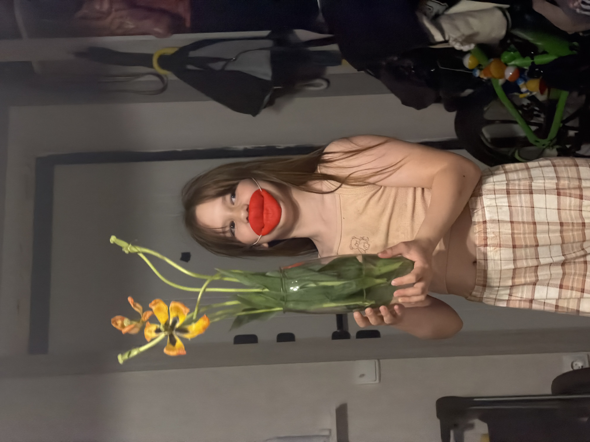 A young girl is holding a vase of tiger lilies. She is wearing a beige tank top and plaid shorts. She has a red oversized lip prop over her mouth. The background shows part of a room interior with objects on shelves. The location is not discernible from the image.
