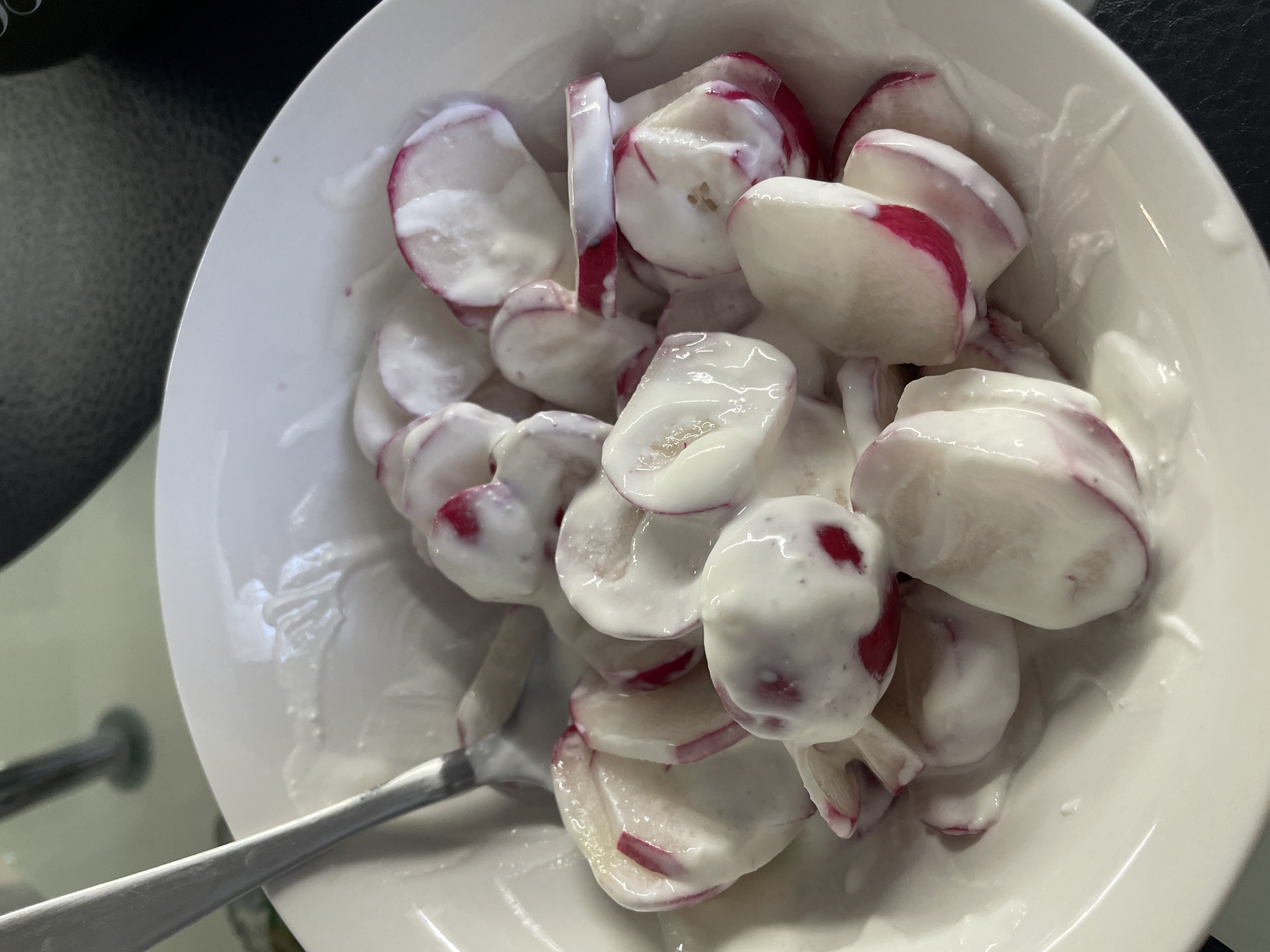 A close-up overhead shot shows a white bowl filled with sliced radishes coated in a creamy white sauce, likely yogurt or sour cream. A silver spoon rests on the left side of the bowl. The scene is brightly lit, highlighting the textures of the food and the glossy surface of the sauce. The background includes a dark, possibly black surface in the upper right and part of what appears to be a chair or other furniture on the left. The exact location in Rozsoshentsi, Ukraine cannot be determined from the visual information.