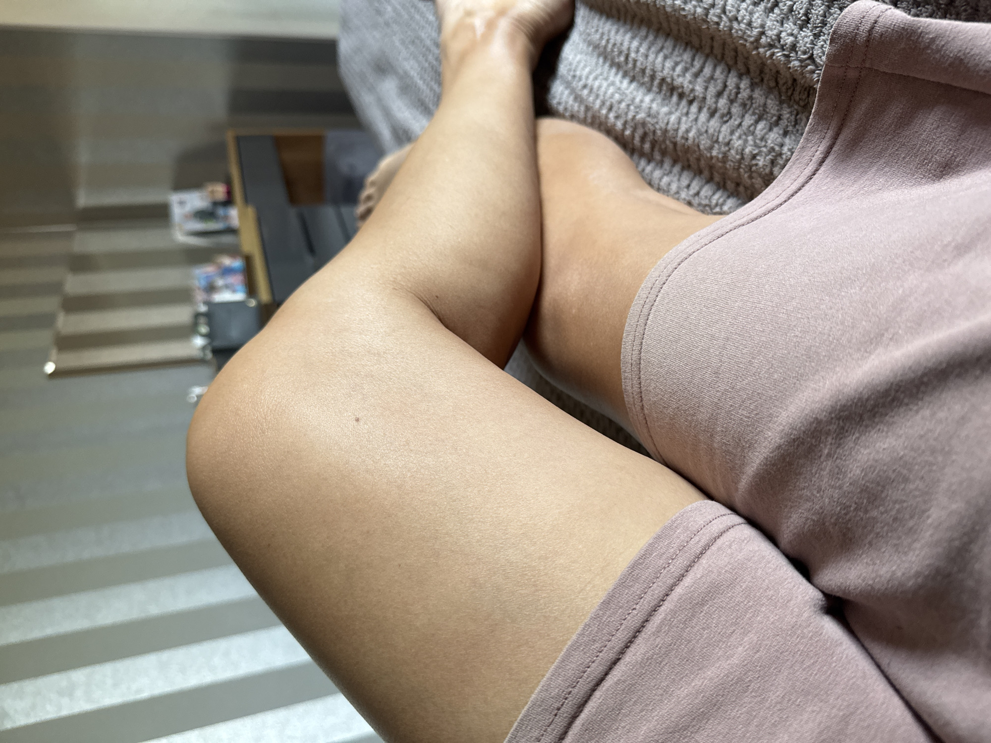 The image shows a close-up view of a person's legs, which are crossed at the knees. The person is lying on a bed with a textured grey blanket. The background of the image includes a dresser with framed pictures on top, and a wall with vertical stripes. The scene is indoors, likely a bedroom. There are no discernible landmarks or features that would specifically place the location in Солонешное, Russia.