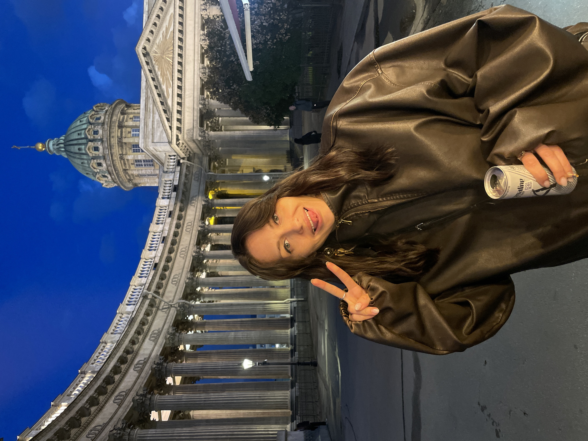 A woman is standing in front of Kazan Cathedral in Saint Petersburg, Russia. She is wearing a brown leather jacket and holding a can of what appears to be a beverage. She is making a peace sign with her right hand and sticking her tongue out. The sky is dark blue, indicating it is nighttime. The cathedral's grand colonnade and dome are visible in the background, illuminated by artificial lights. The street is paved, and there are streetlights and some trees visible.