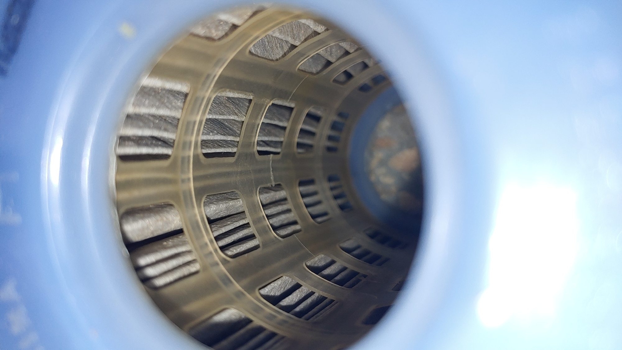 The image is a close-up, slightly blurry, interior view of a blue cylindrical object. The object appears to be a container or housing, with a blue plastic rim visible at the top and bottom. Inside, there is a filter element composed of pleated gray material supported by a translucent beige plastic cage. The cage has a pattern of interconnected rectangular openings. The pleated filter material is visible through these openings, showing a tightly folded, dark gray texture. The background within the cylinder is dark and out of focus, with some speckled brown and gray tones. There are no people or discernible actions in the image. The provided context of "Mukachevo, Ukraine" is not visually supported by the image content.