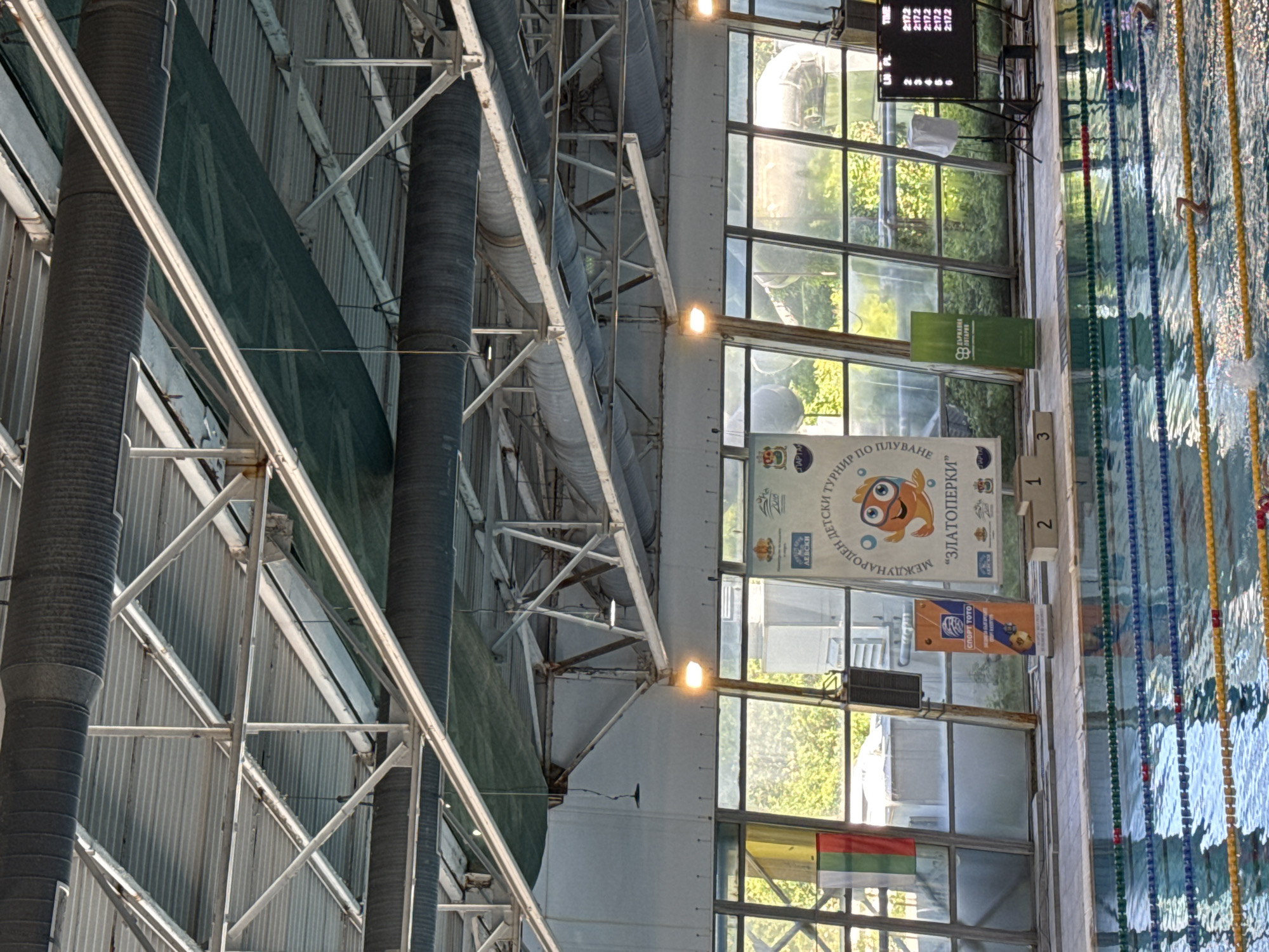 Here is a description of the image:
The image shows the interior of a swimming pool facility. Swimming lanes are visible in the pool water on the right side of the image. There are starting blocks with numbers "1", "2", and "3" marked on them. Above the pool area, a series of windows allows natural light into the building. Banners and flags are suspended from the building structure, including a banner for the "Zlatoperki" International Children's Swimming Tournament and the Bulgarian flag. An electronic scoreboard showing times is located on the left of the pool. The ceiling of the pool features a complex metal framework with pipes and ventilation systems.