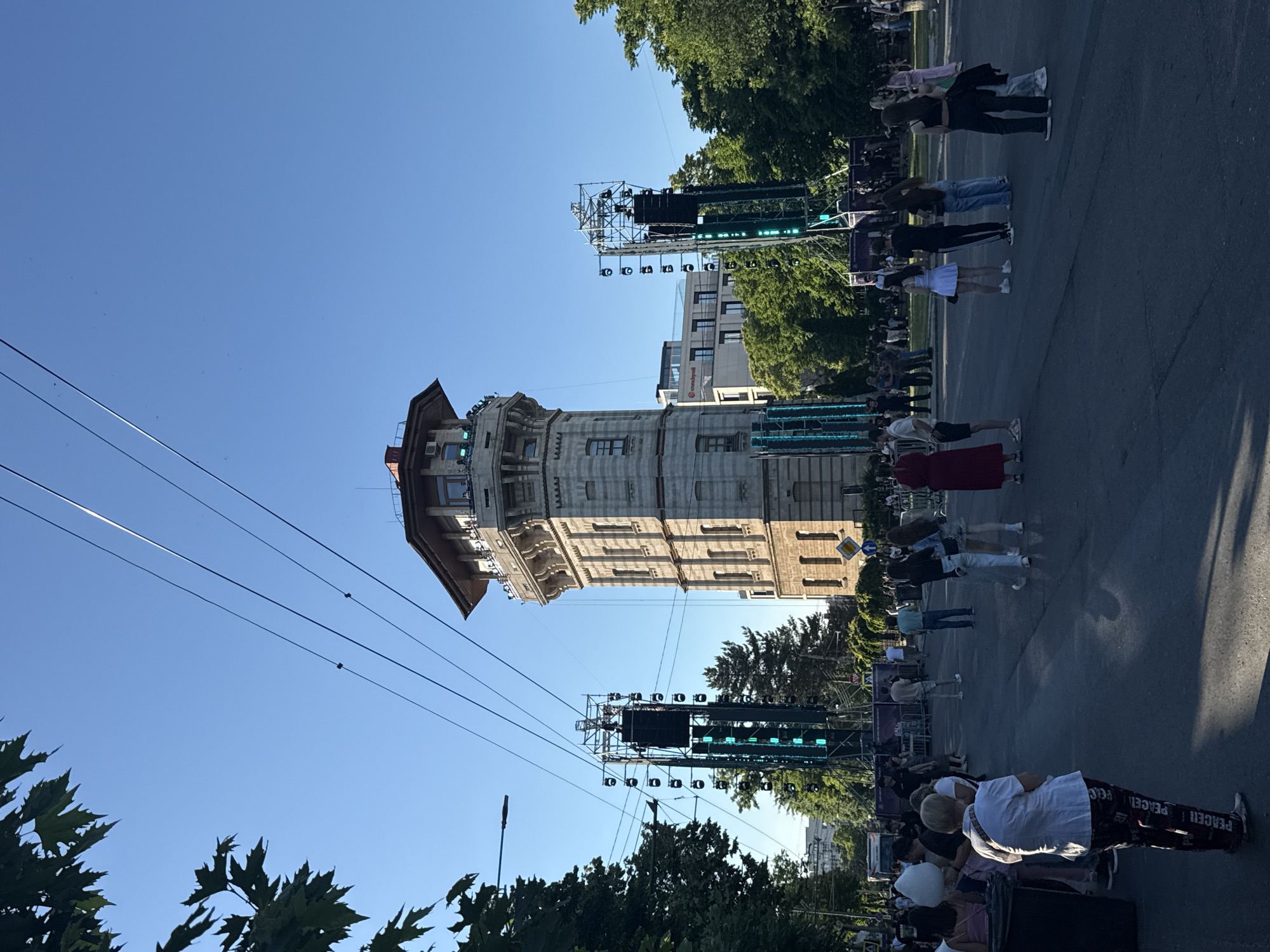 The image depicts an outdoor event with a tower in the background, identified as the Water Tower in Chișinău, Moldova. Several large speaker systems and lighting rigs are set up on metal scaffolding, indicating a concert or festival. A crowd of people is gathered on a paved area in front of the tower and stage structures, engaged in various activities such as walking, standing, and interacting. The sky is clear and blue, suggesting daytime. Trees line the sides of the area, and buildings can be seen behind the tower. The overall scene captures a lively public gathering in an urban setting.