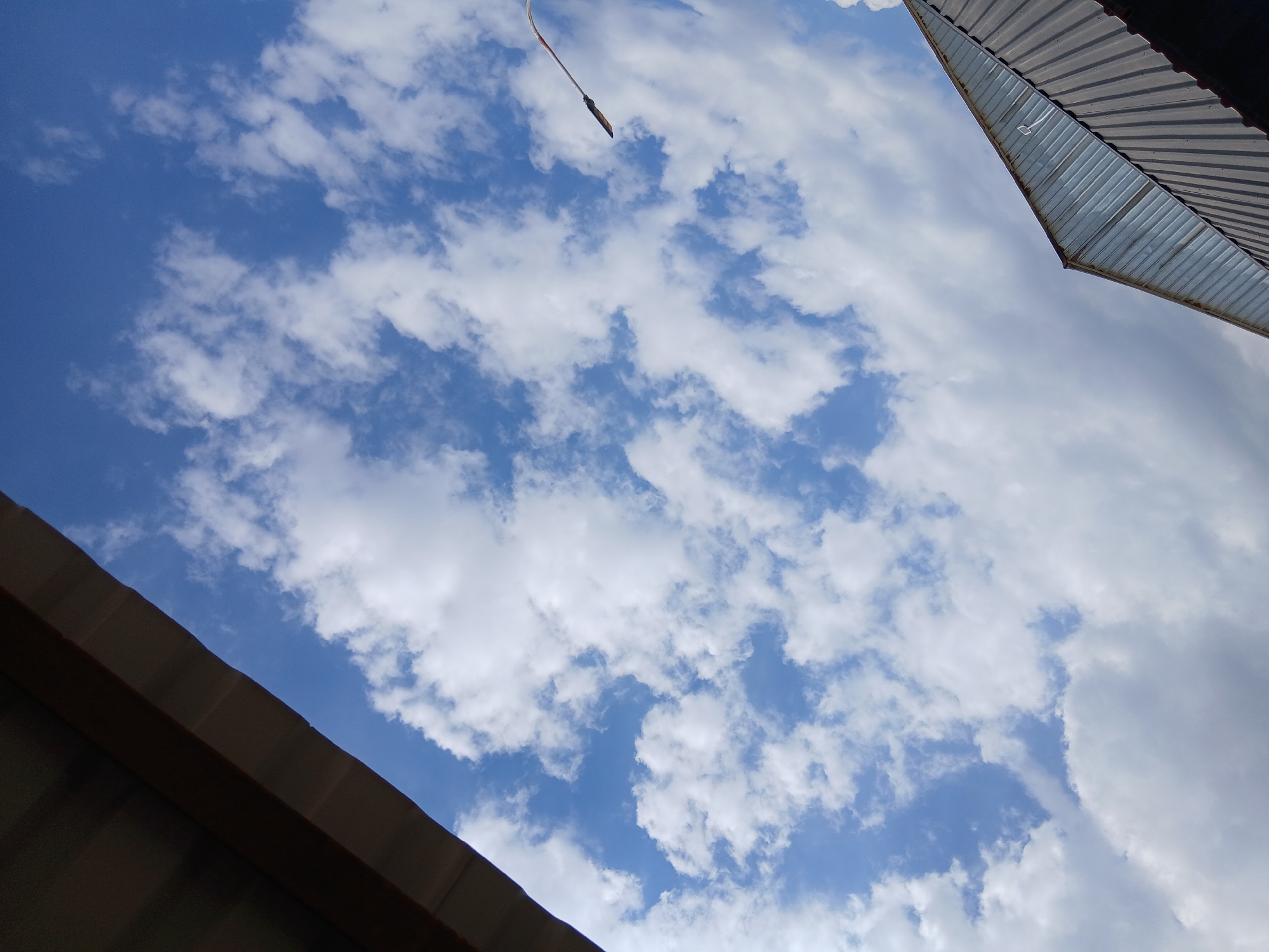 The image shows a view looking up at a blue sky with scattered white clouds. Two parts of a building's roof are visible in the foreground, one in the upper left corner and another in the lower right. The roof in the upper left appears to be dark and corrugated. The roof in the lower right is also corrugated and appears to have sections of glass or translucent material. A portion of a street lamp or utility pole is visible in the upper right quadrant of the image, extending diagonally upwards. The scene is outdoors, and the sky suggests daytime. The location is identified as being in the city of Kanchoch, Tajikistan.
