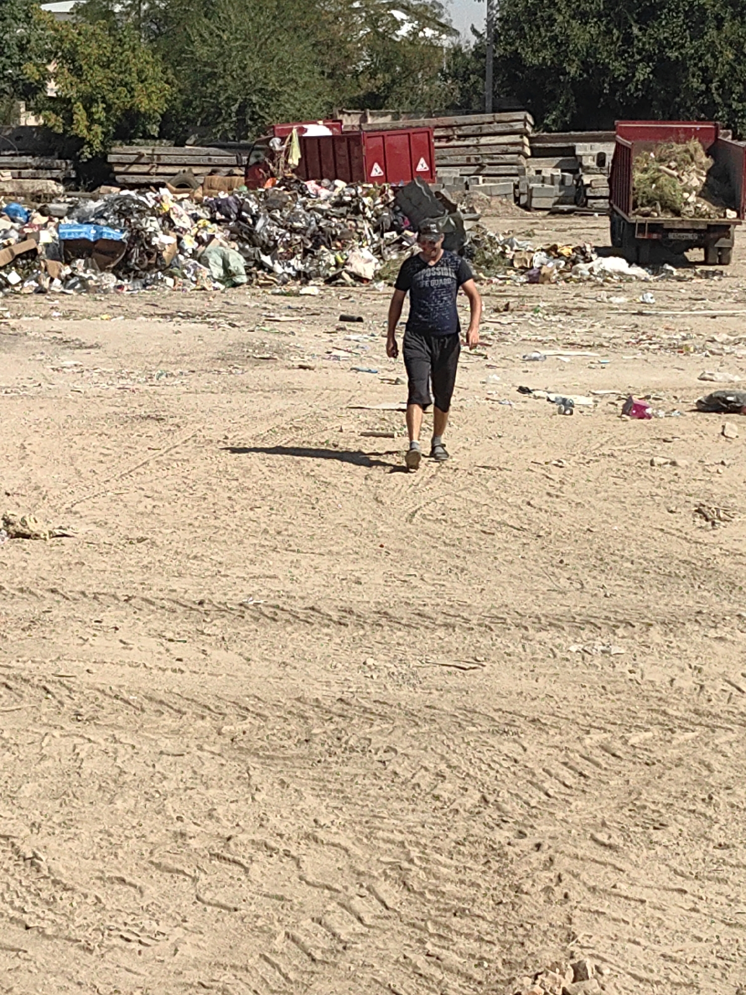 The image depicts a landfill site in the city of Shymkent, Kazakhstan. A person is walking across the sandy, debris-strewn ground. In the background, there are large piles of garbage, dump trucks, and stacks of construction materials. The environment appears to be an open, outdoor area with trees in the distance. The overall scene suggests a waste disposal or recycling facility.