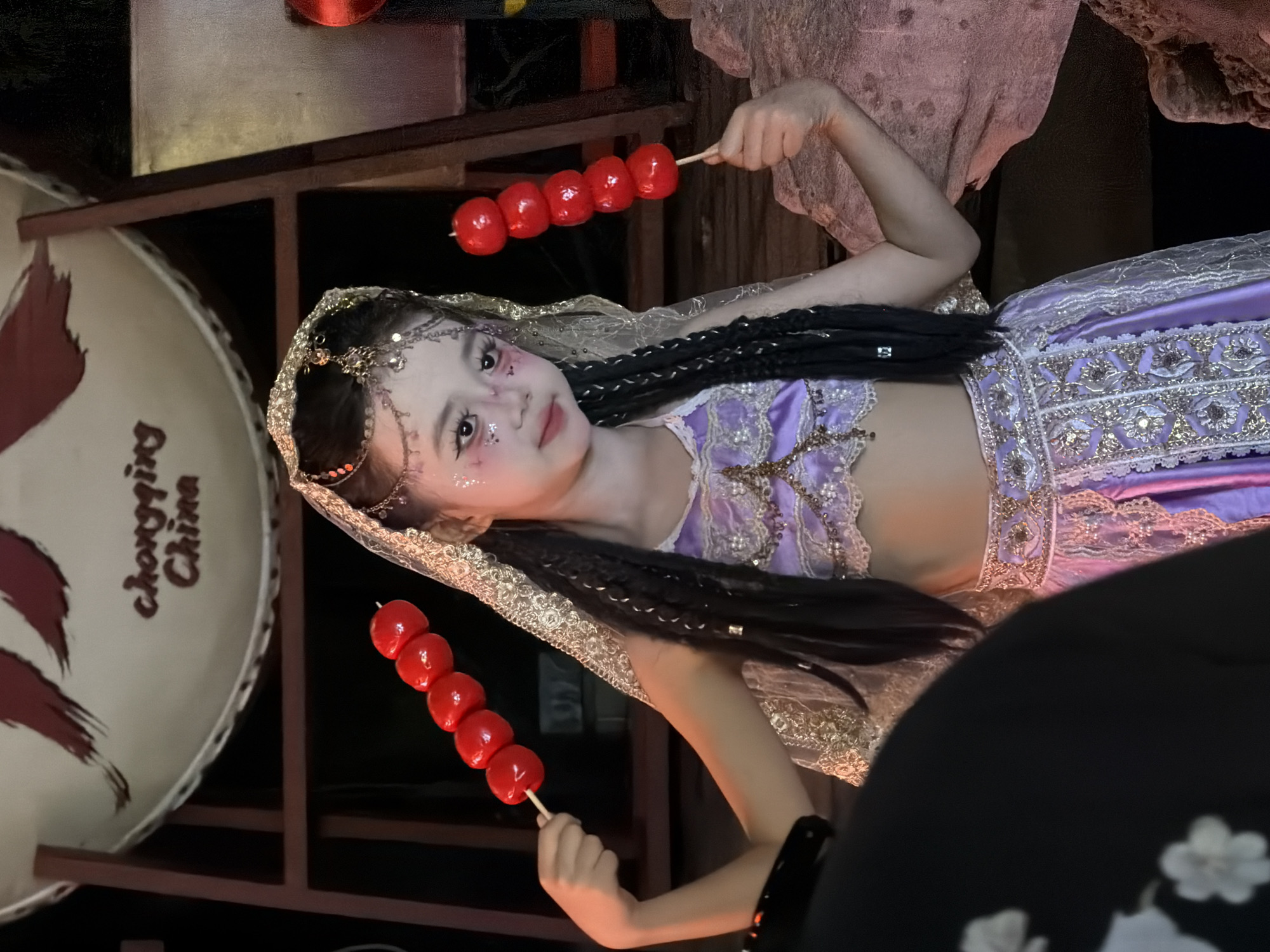 A young girl, adorned in a traditional purple and gold outfit and intricate headwear, poses for a photo. She holds a skewer of candied red fruit in each hand, bringing one close to her face. Her eyes are looking towards the camera with a gentle smile. Her hair is styled in long, dark braids that fall over her shoulders and down her back. The background is dimly lit, suggesting an indoor setting or evening time. A large, circular object with faded red markings and the partial text "chongqing China" is visible on the left side of the image. The girl's attire and the presence of traditional elements suggest a cultural performance or celebration.