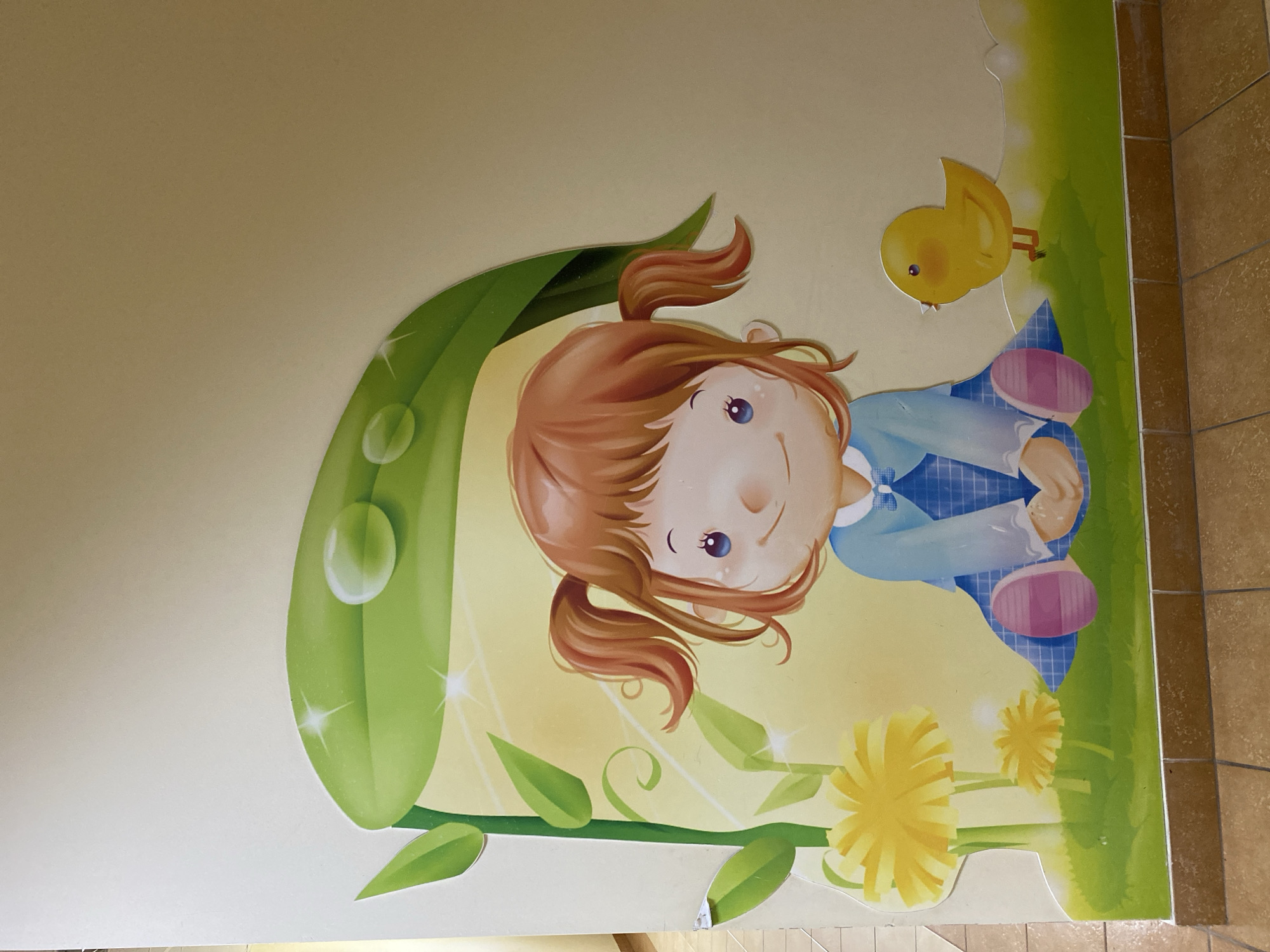 The image shows a mural on a wall, depicting a young girl with auburn hair in pigtails, wearing a blue dress with a plaid pattern and pink shoes. She is sitting on green grass with yellow flowers. Above her head, a large green leaf with water droplets acts as a canopy. To her right, a small yellow chick is standing. The mural is colorful and has a childlike aesthetic, suggesting it is likely located in a place intended for children, such as a kindergarten, daycare, or a children's play area. There is no discernible information in the image that can be used to determine its location in the city of Kaliningrad, Russia. The scene is static and does not depict any actions or interactions between the depicted characters.