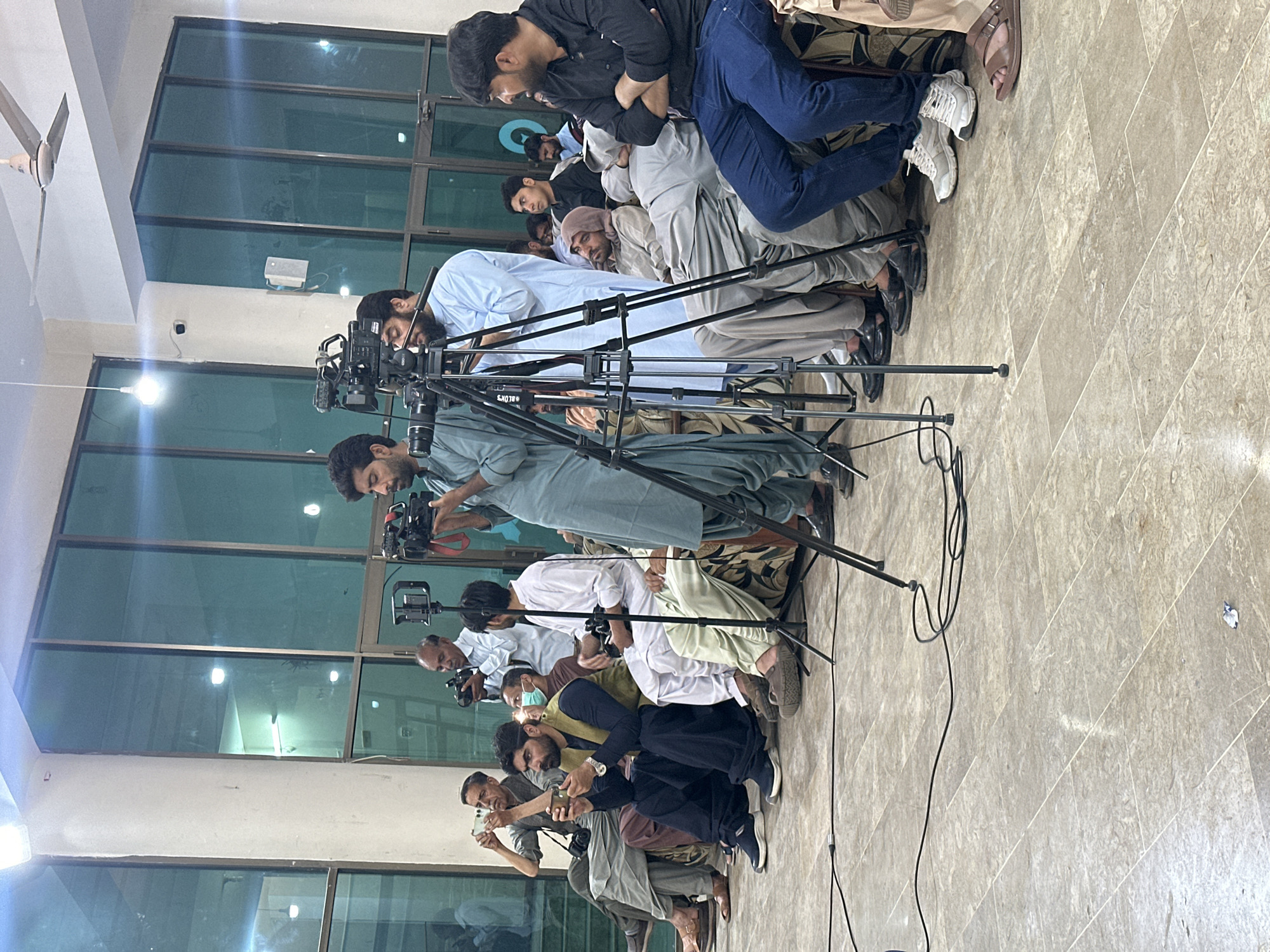 A line of men sits on a patterned floor in what appears to be a media event in Quetta City Tehsil, Pakistan.
The men are facing a wall, some holding and operating cameras and phones. The first few men in the line are using professional cameras mounted on tripods. Other individuals further down the line are using phones to take pictures. All are aimed in the same direction. The room has a reflective surface of windows or glass, reflecting the indoor lighting. The building appears to be modern with a ceiling fan visible. The focus is on the photographers and their equipment. Cables from the cameras are visible on the floor. The men wear various styles of clothing. Some have traditional attire.