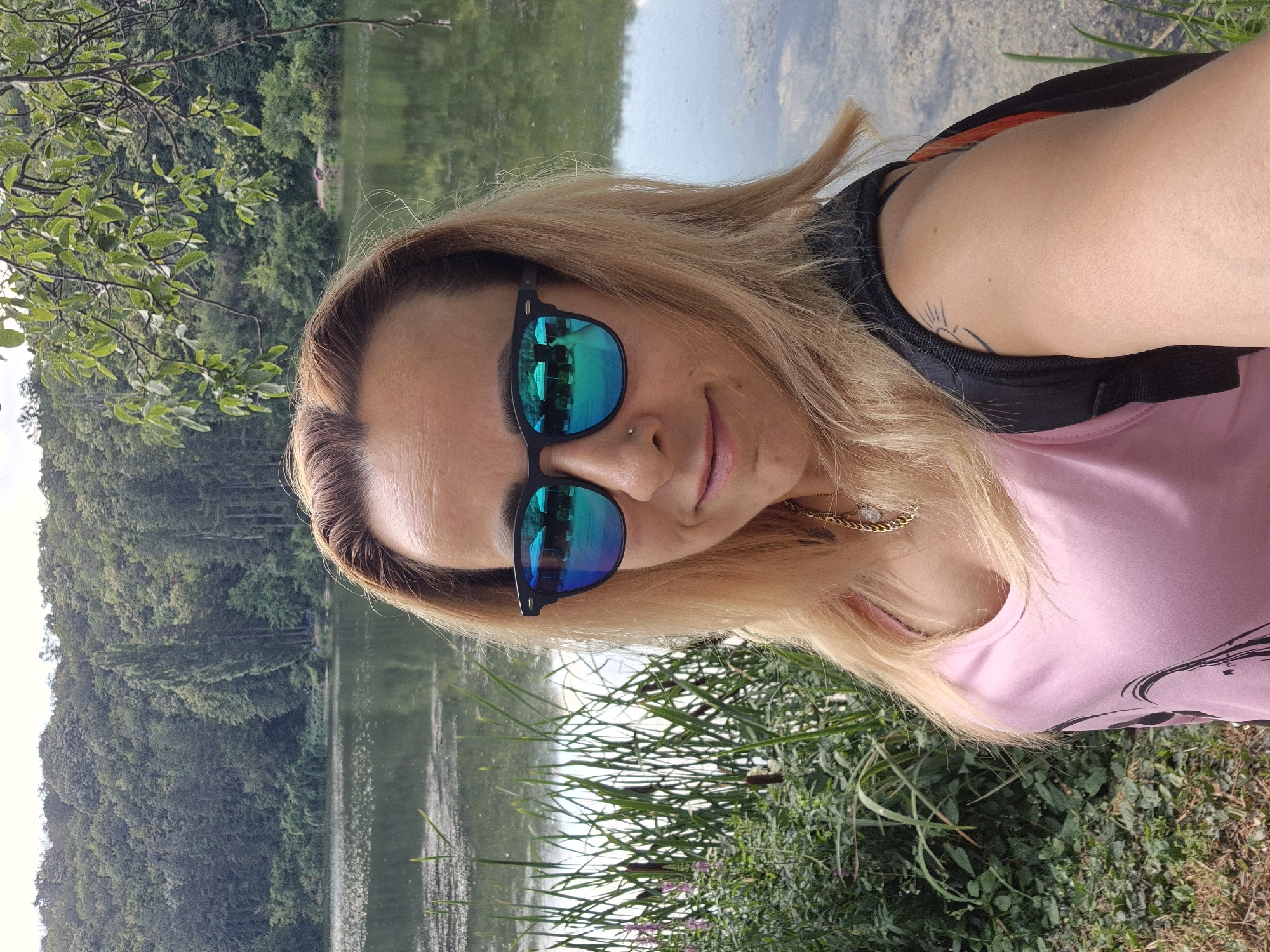 A medium shot captures a woman with blonde hair and sunglasses taking a selfie by a lake. She is wearing a pink tank top and a backpack. The background shows a forest and the lake. The scene appears to be outdoors in a natural setting, possibly a park or a rural area.