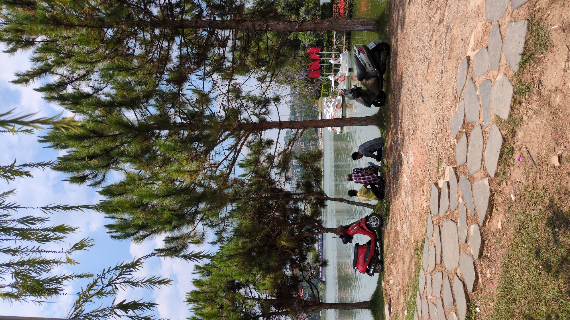 The image depicts a park scene in Da Lat, Vietnam, specifically near Xuan Huong Lake. Three people are crouched at the edge of the lake, near two motorbikes, one red and one silver. The setting includes a paved path, trees, and a view of the lake with decorative boats. In the background, buildings and foliage are visible across the lake.