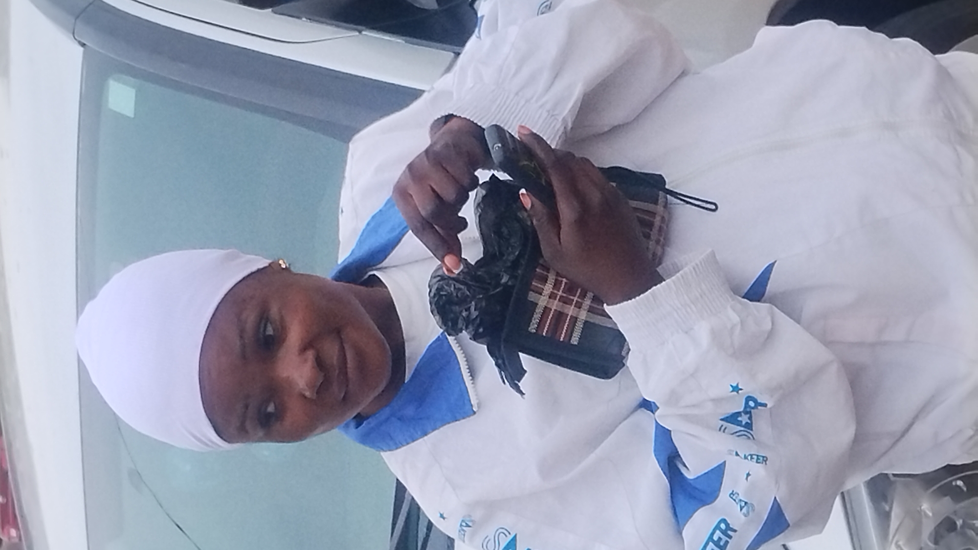 A woman wearing a white headscarf and a white and blue jacket stands in front of a white vehicle. She is holding a dark, possibly black, object in her hands. The object appears to be wrapped in a checkered cloth. Her left hand is holding the object, and her right index finger is touching it. The woman has a neutral expression. The background is blurred, but the vehicle suggests an outdoor urban setting. The image is taken from a medium distance, showing the woman from the chest up. It is not possible to determine the exact location within the city of Jos, Nigeria, from the provided image.