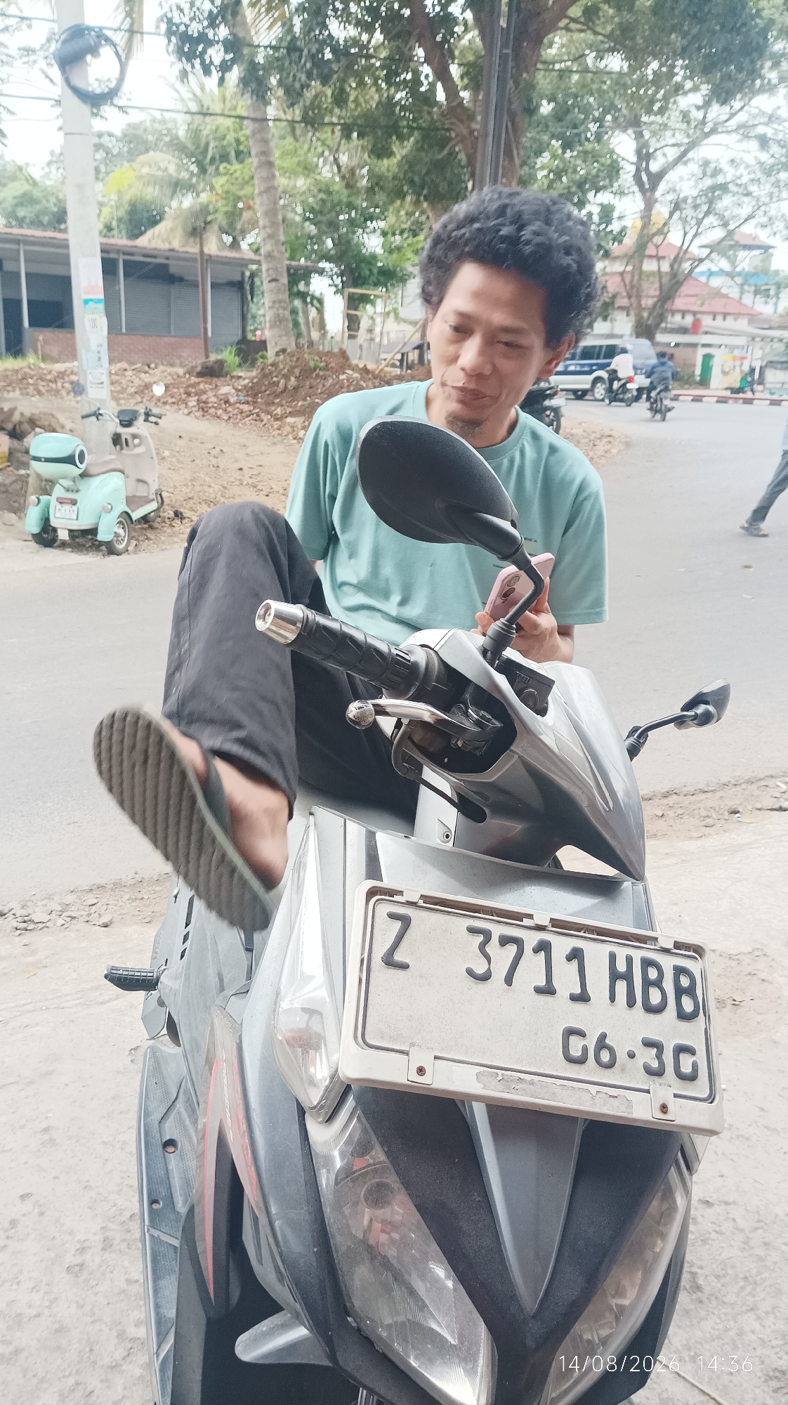 A man with curly dark hair and a teal t-shirt is sitting on a motorcycle, holding a pink smartphone. He is wearing black pants and flip-flops. The motorcycle has a license plate that reads "Z 3711 HBB C6-3C". In the background, there is a light blue scooter, trees, buildings, and other vehicles. A timestamp on the image reads "14/08/2026 14:36". The scene appears to be an outdoor urban environment.