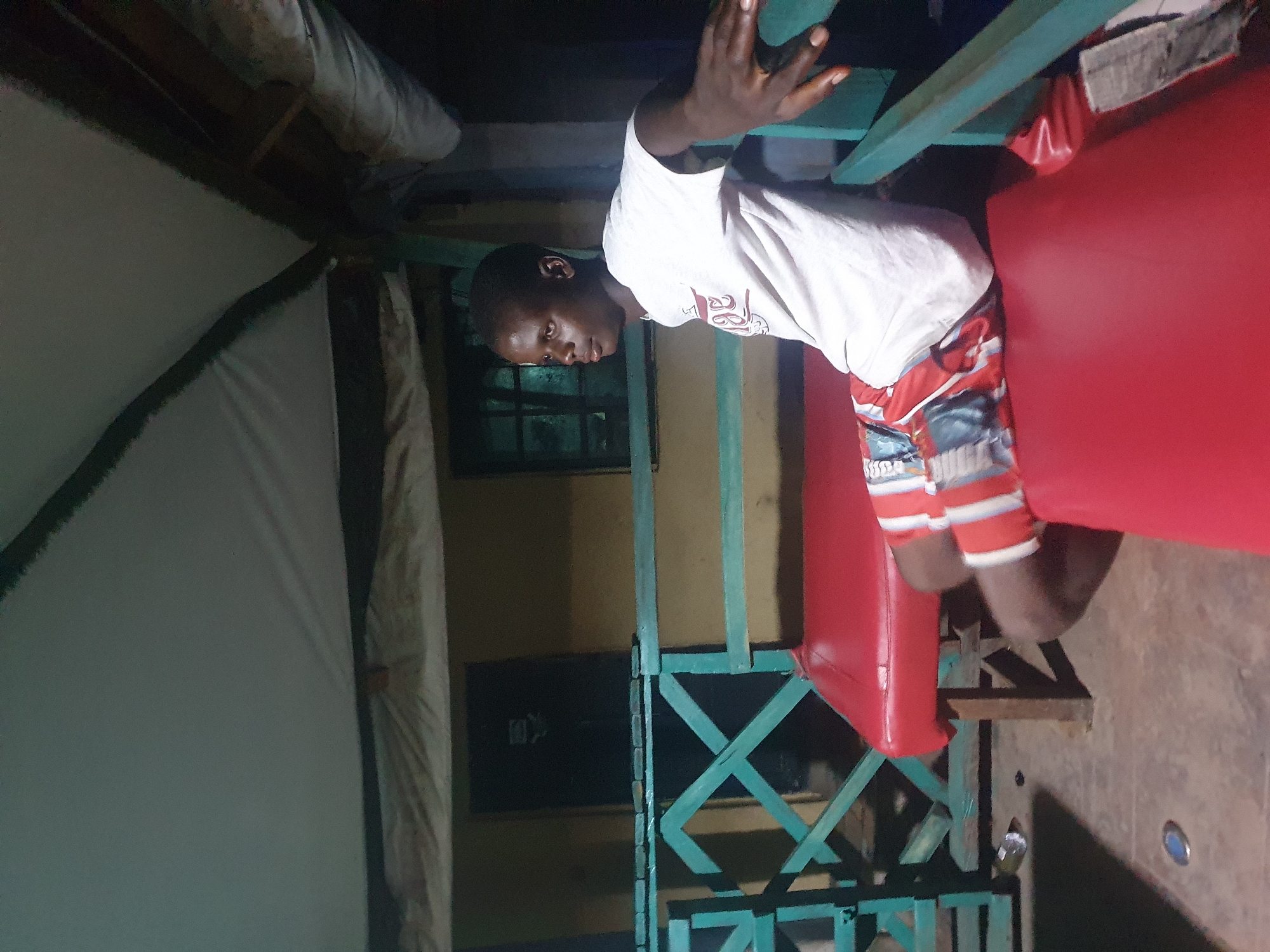 In Lokoja, Nigeria, a young Black male is seated on a red bench under a tent-like structure. He is wearing a white t-shirt with a graphic design on the front and red and white striped shorts. The man is looking to his right, with his right arm resting on the green railing of the bench. The bench has a wooden base with red upholstery. Behind the bench is a wall with a window. The ground is made of concrete and is sparsely littered with a few items, including a discarded can. The overall lighting suggests it is either early morning or evening, with the tent providing shade.