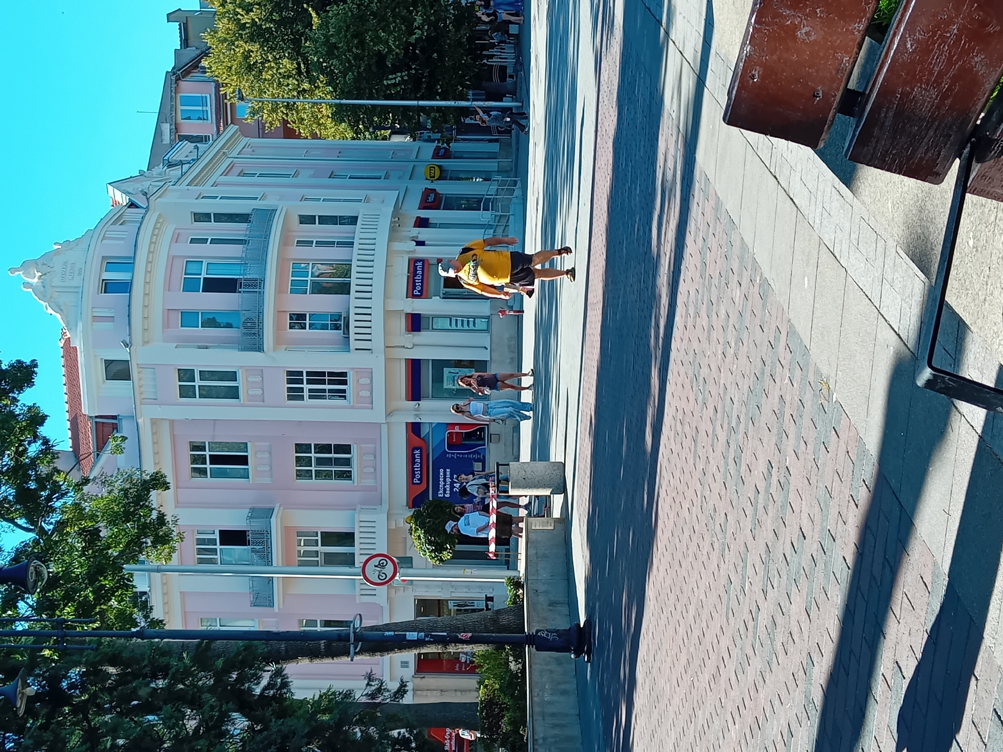 The image depicts a sunny street scene in Varna, Bulgaria. In the foreground, a brick-paved walkway is visible, with shadows cast by trees and possibly overhead structures. A dark brown wooden bench is positioned in the lower right corner of the frame.
Further back, a light pink building with white trim and balconies dominates the scene. The building has multiple windows and appears to be a commercial establishment with a "Postbank" sign clearly visible. In front of the building, a group of people are walking and standing. One individual, a man wearing a yellow t-shirt and shorts, is walking towards the right of the frame, carrying a backpack. Several other people, including women in casual attire, are also present in the vicinity of the Postbank.
A lamppost and a bicycle prohibition sign are also visible in the middle ground. Trees with green foliage provide a natural element to the urban environment. The overall atmosphere is one of a bustling city street on a bright day.