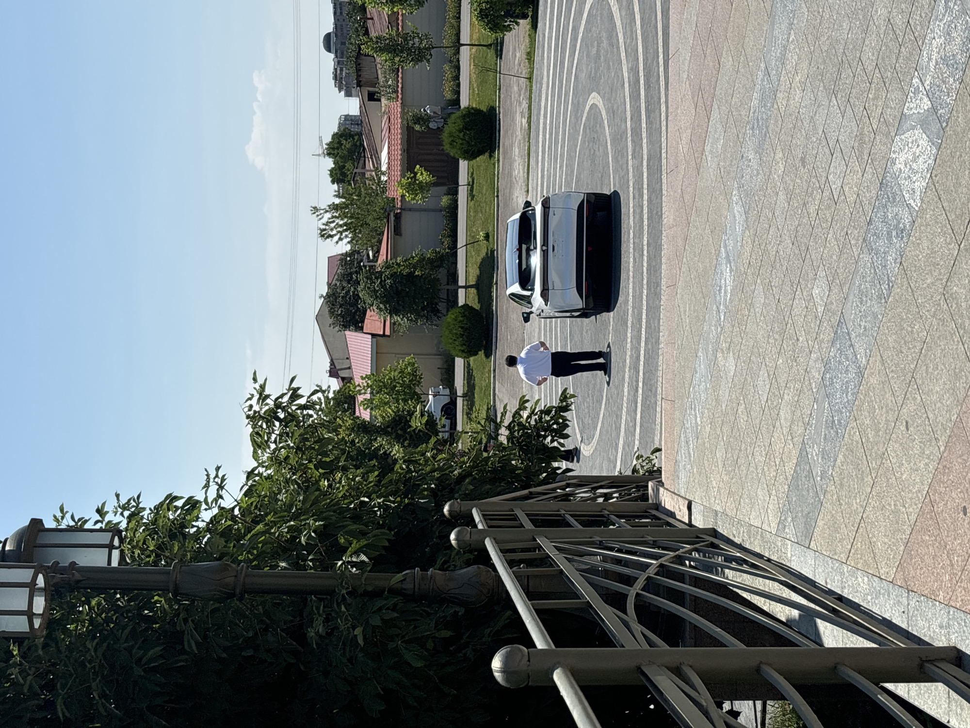 A man in a white shirt and dark pants stands facing away from the camera, near the rear of a white electric car parked in a circular paved area. The car has a black roof and tinted windows. The paved area has a pattern of concentric circles and segmented lines. The scene appears to be outdoors in a city, likely Tashkent, Uzbekistan, given the architectural style of the buildings visible in the background, which include red-tiled roofs and stucco walls. A decorative lamppost with a glass lantern is positioned to the left of the frame, partially obscured by lush green foliage. A metal railing runs along the left side of the foreground, suggesting a raised walkway or stairs. The sky is clear and blue.