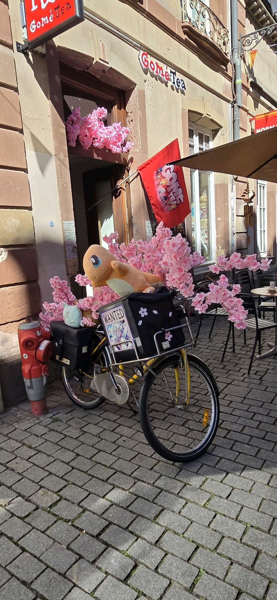 This is a medium shot, eye-level, outdoor photograph of a yellow bicycle parked on a cobblestone street in Strasbourg, France. The bicycle is decorated with pink artificial cherry blossoms and has a black storage bag on the back. A plush orange Charmander toy is placed on top of the bag, and a light blue plush toy is sitting on the left side of the bag. A "WANTED" poster featuring a character illustration is attached to the storage bag. A red fire hydrant is located to the left of the bicycle. In the background, a building with brown stone facade has a store sign that reads "Gome Tea". There are also outdoor tables and chairs visible in the background, suggesting a cafe or restaurant setting. The overall scene is bright and sunny.