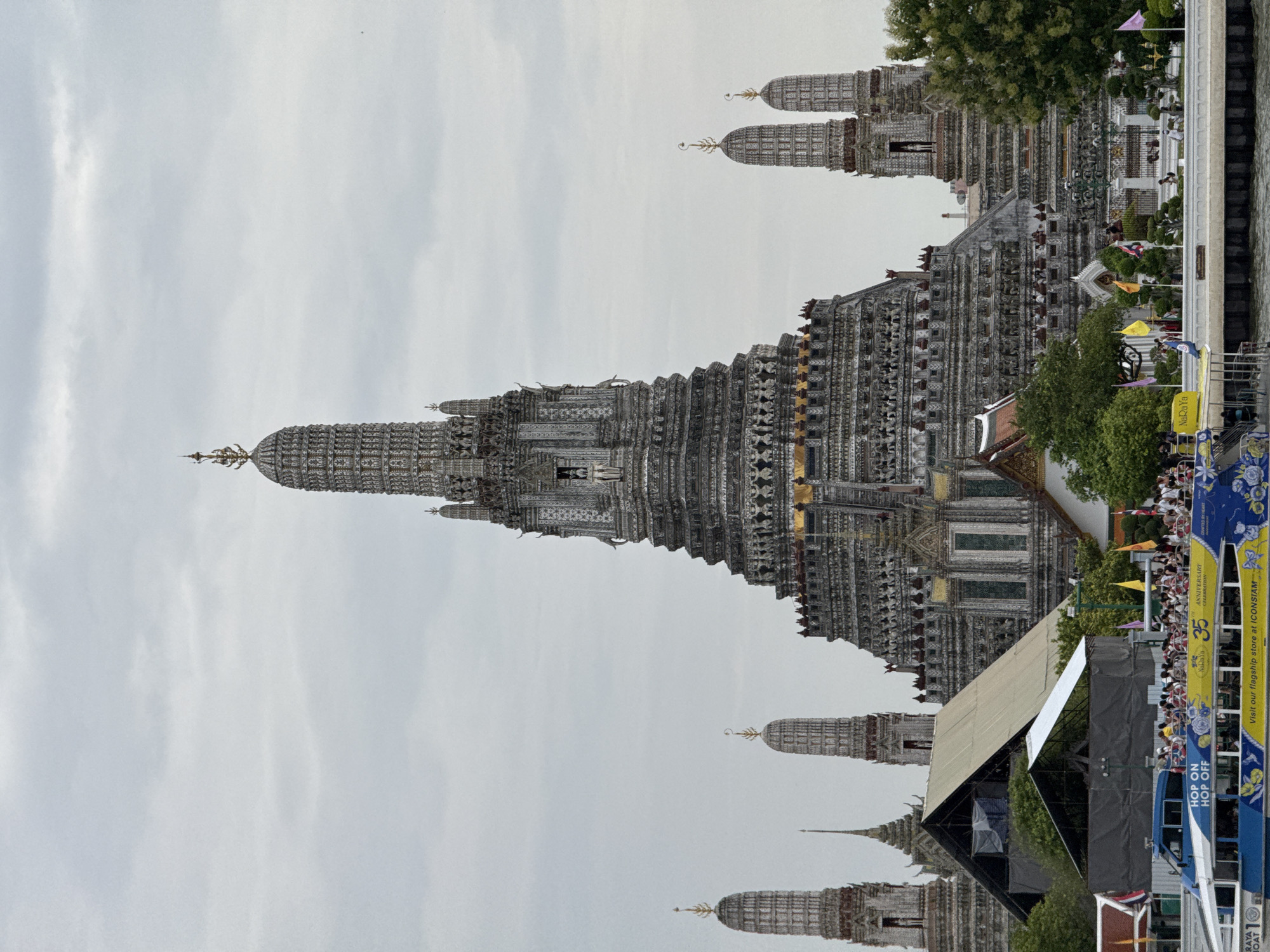 The image captures the Wat Arun (Temple of Dawn) in Bangkok, Thailand, viewed from the Chao Phraya River. The central and largest structure is the main prang, a towering spired temple adorned with intricate white porcelain and detailed carvings. Flanking the main prang are several smaller prangs of similar architectural style. In the foreground, a blue and yellow ferry boat is laden with tourists. The boat has text indicating "HOP ON HOP OFF" and "NaRaYa 35 ANNIVERSARY CELEBRATION". People are visible on the deck of the boat, and some are standing along the riverbank. Lush green trees and parts of other temple structures are visible behind the riverbank. The sky is overcast and grey.