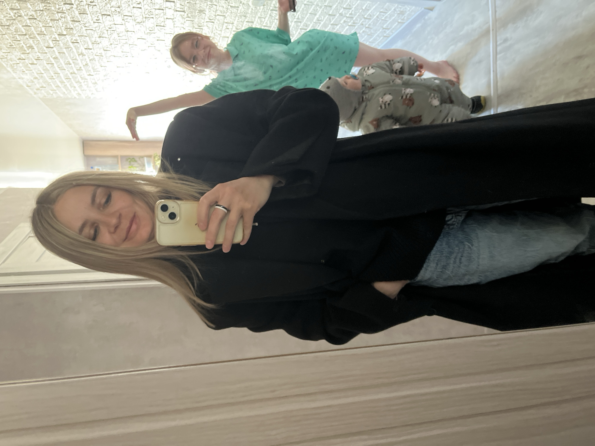 This indoor mirror selfie, likely taken in Chaikovsky, Russia, captures three individuals reflected in what appears to be a large wall mirror.
In the foreground, a woman with long blonde hair is taking the selfie, holding a light-colored smartphone with her left hand. She wears a black top or cardigan and light blue jeans, and a black and white ring is visible on her left ring finger. She has a subtle, gentle smile on her face.
In the background, visible as a reflection above the selfie-taker's shoulder, are two other individuals. Another woman with light brown hair, tied up, is smiling broadly. She wears a light green short-sleeved dress with a repeating pattern, possibly small light-colored shapes. One of her arms is extended, as if posing or reaching. Next to her, a young child is lying or crawling on the floor, reflected upside down from the perspective of the mirror. The child is dressed in a grey or greenish full-body onesie patterned with animal figures, such as bears. A pacifier is visible near the child's mouth.
The setting appears to be a brightly lit interior room, possibly with a textured white ceiling and light-colored walls and floor. The overall mood conveyed by the smiles is cheerful and relaxed. There is no visible text in the image.