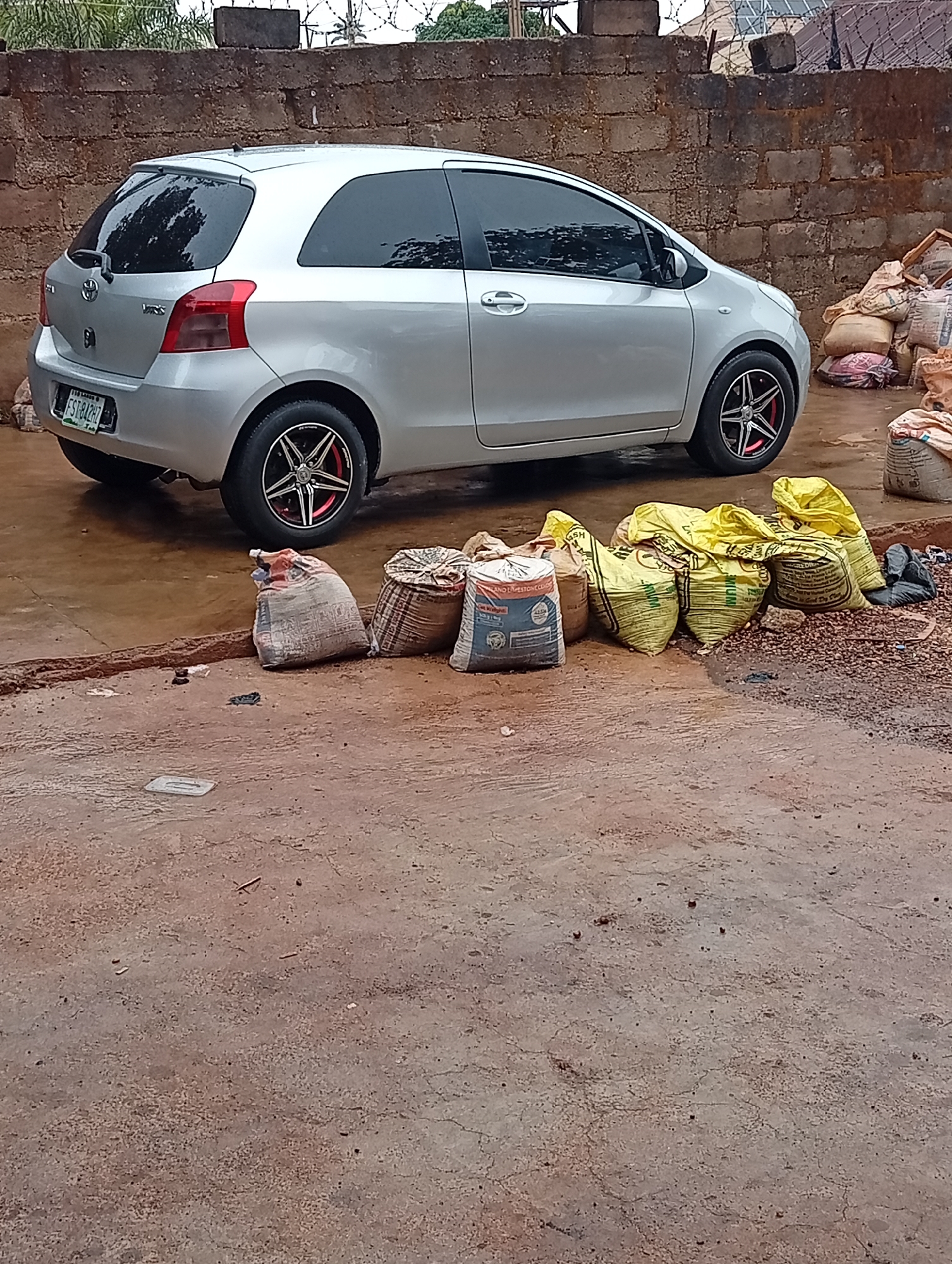 A silver Toyota Yaris is parked on a concrete surface next to a collection of sacks and construction materials. The car is positioned in front of a brick wall. The environment appears to be a semi-urban or rural area, possibly in Gura Top, Nigeria. The scene appears to be outside and possibly on a construction site or storage area. There are no people visible.