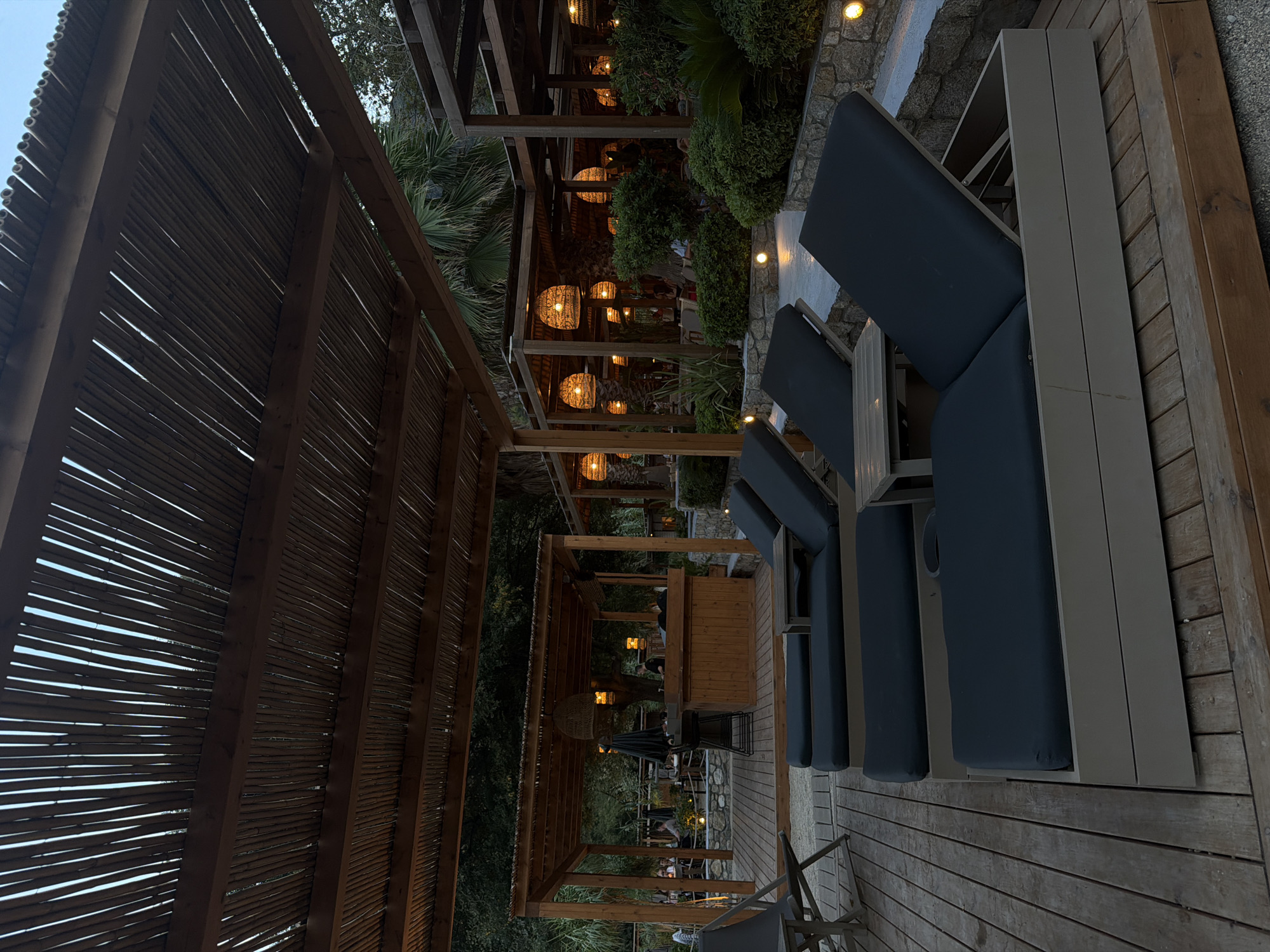 The image depicts an outdoor seating area with lounge chairs on a wooden deck in Sithonia Municipal Unit, Greece. In the foreground, several dark-colored lounge chairs are arranged on a weathered wooden platform. Behind the chairs, a wooden structure serves as a bar area, with a bartender visible. Further in the background, more seating areas with tables and chairs are visible, illuminated by woven pendant lights. Lush green foliage and palm trees surround the area, suggesting a tropical or Mediterranean setting. The overall atmosphere is relaxed and invites patrons to unwind.