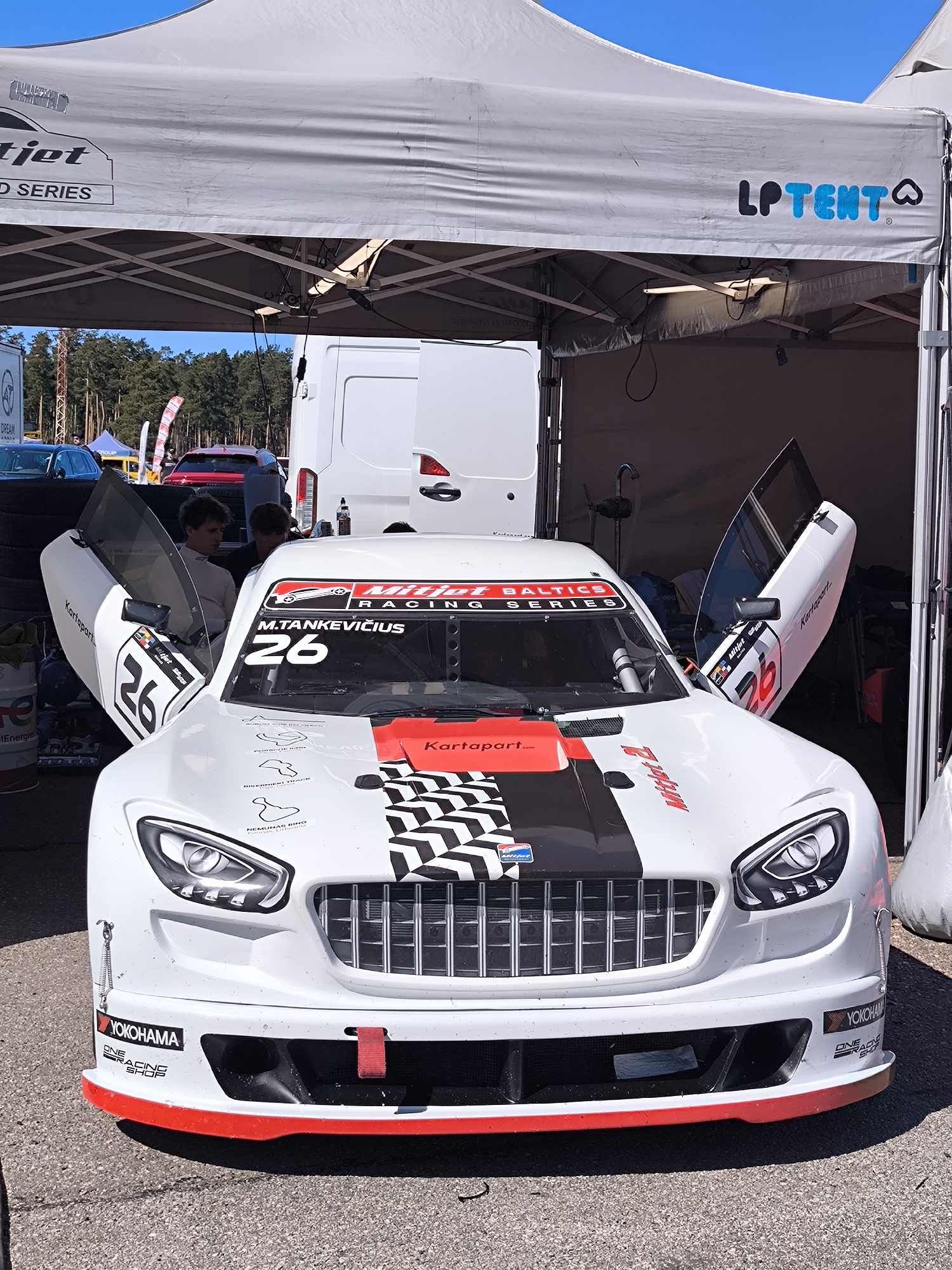 A white Mitjet BALTICS Racing Series car, numbered 26, is parked under an LP Tent. The car has its doors open, revealing two people inside. The car has various logos and decals on it, including 'Kartapart', 'Mitjet', 'Yokohama', and a graphic depicting various racetrack layouts. The backdrop features a white van and trees.