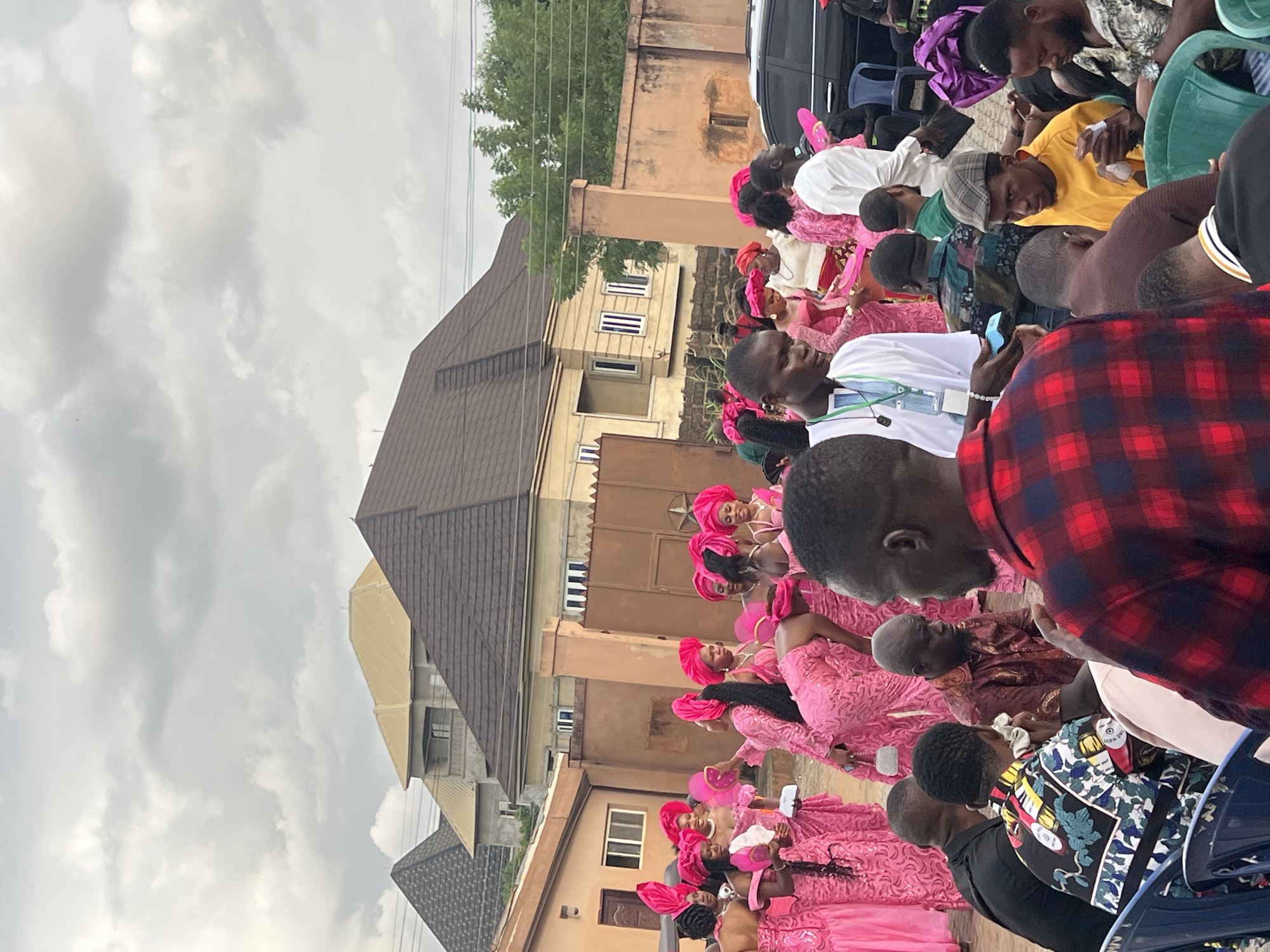 A wide shot depicts a gathering of people, presumably at a wedding ceremony. Many are dressed in matching pink outfits and head coverings. Others wear a variety of casual clothing. The setting is outdoors, next to buildings, under a cloudy sky.