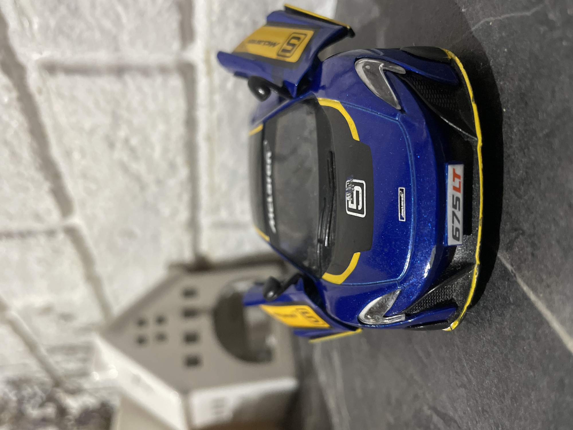 A blue and yellow toy McLaren 675LT is displayed on a dark, textured surface, possibly a countertop or shelf. The toy car is positioned front and center, with its dihedral doors open. The backdrop features a white brick wall and a miniature white house with arched doorways and small square windows. The overall scene is indoors, likely in a domestic setting. There is no indication of the city of Baranovka, Russia, in the image.