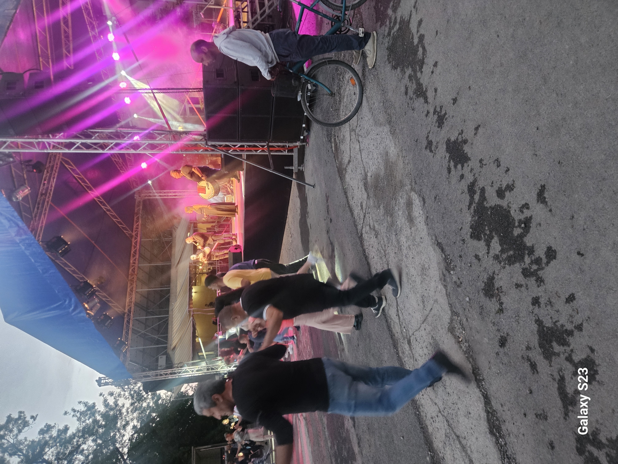 The image shows an outdoor concert stage in Sliven, Bulgaria. The stage features a steel truss structure with lighting, including pink spotlights. A band is performing on stage. A crowd of people is watching the performance. In the foreground, several people are visible from the waist up, facing the stage. One man is standing next to the stage with a bicycle. The ground is asphalt with dark spots. "Galaxy S23" is printed on the right side of the image.