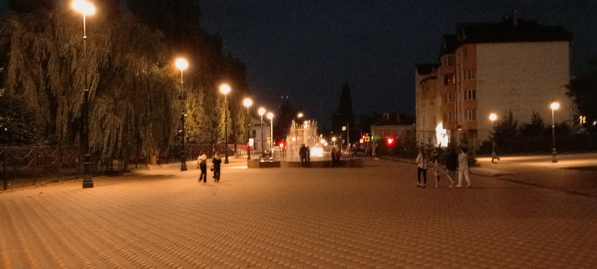The image depicts a nighttime scene in Kirsanov, Russia, specifically along a paved pedestrian walkway illuminated by streetlights. The walkway is made of light-colored cobblestones laid in a herringbone pattern. On the left side of the walkway, a row of trees with cascading branches, possibly weeping willows, are situated behind a decorative metal fence. Several streetlights are visible along this side, casting a warm glow.
In the mid-ground, a fountain with illuminated water jets is present. A group of four individuals, two adults and two children, are walking towards the fountain from the right side of the image. To the left of the fountain, a couple or two individuals are walking away from the viewer. Further down the walkway, near the fountain, there are more figures, their details blurred by the distance and low light. Traffic lights are visible on poles, showing red and green signals.
On the right side of the walkway, a multi-story building with cream-colored walls and multiple windows is prominent. Another building, likely residential, with orange and cream-colored sections and dark windows, is situated behind it. Several more streetlights are spaced along the right side of the walkway, illuminating the area. The sky is dark, indicating nighttime. The overall atmosphere is one of an urban public space after dark, with people enjoying the illuminated fountain and promenade.