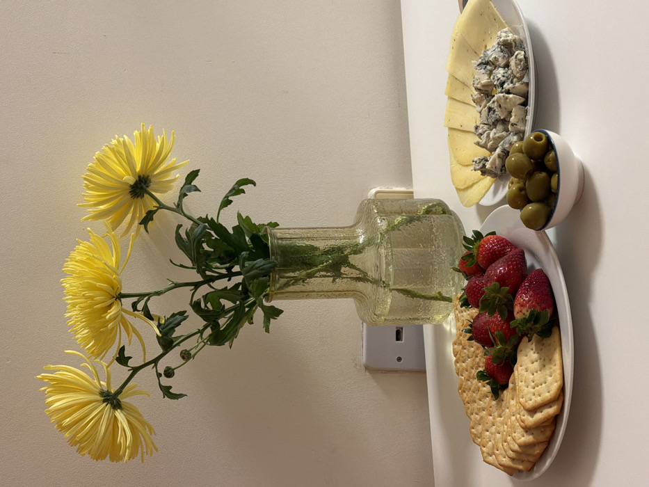 This image displays an indoor scene featuring a table arrangement against a light-colored wall. The specific location in Tel-Aviv, Israel, is not discernible from the visual content.
Central to the composition, a clear glass vase containing water holds three large yellow spider chrysanthemums and green foliage, including one visible bud. Behind the vase, a white two-pin electrical socket is mounted on the wall.
On the white table surface in the foreground, two white round plates and one small white bowl are present. The plate on the left contains an arrangement of plain crackers, some round and some rectangular, alongside approximately ten fresh red strawberries with green calyxes. The plate on the right holds several slices of yellow cheese and a quantity of crumbled blue cheese. Between these two plates, a small white bowl with a blue rim is filled with green olives. No people are visible, and no actions or interactions are depicted.