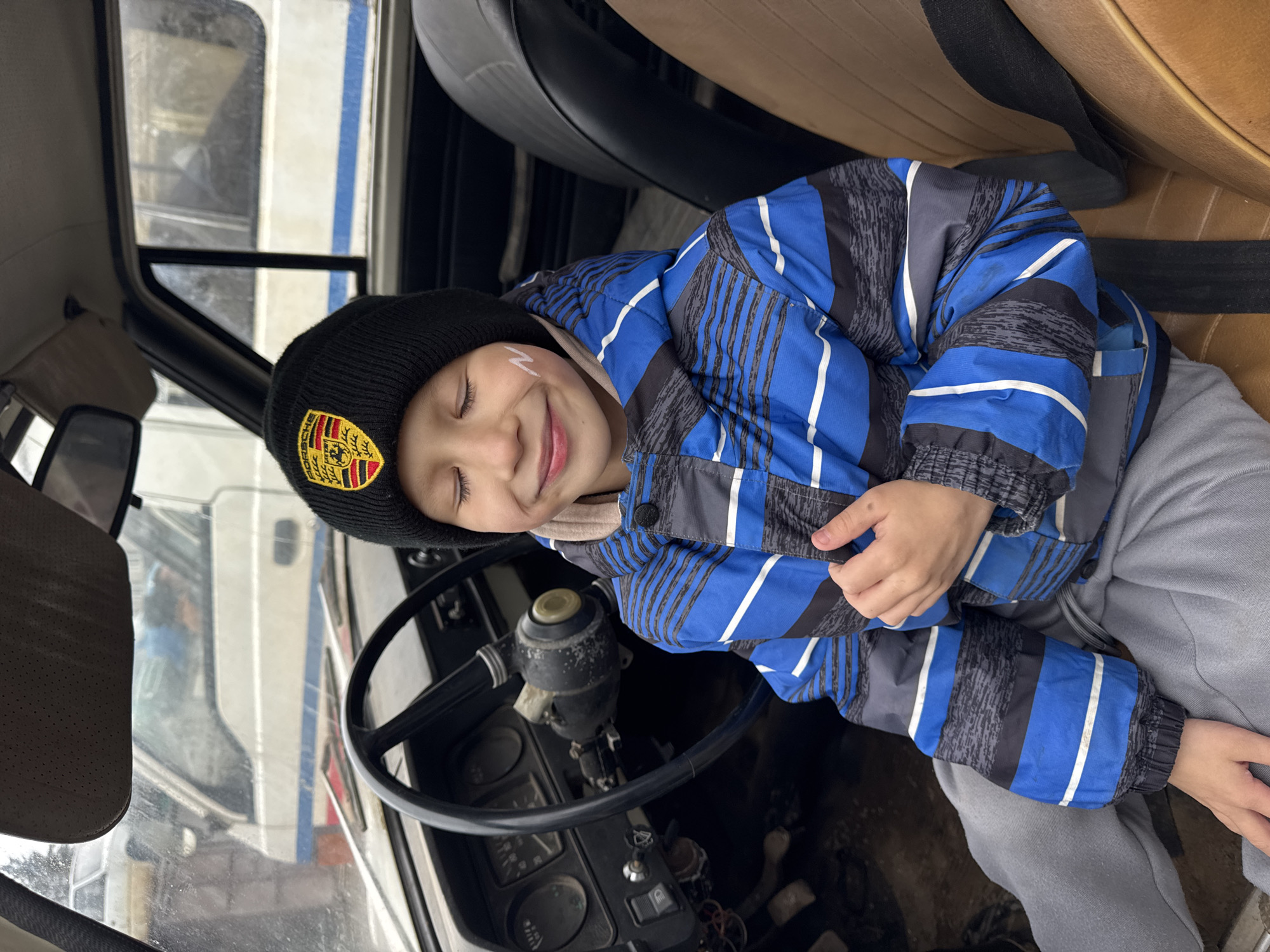 A young boy is sitting in the driver's seat of an old vehicle. He is wearing a black beanie with a Porsche emblem and a blue and grey striped jacket. His eyes are closed, and he has a small, white painted design on his cheek. The vehicle's steering wheel and dashboard are visible. A seatbelt is wrapped around the boy. The location is Chaikovsky, Russia. The precise location within Chaikovsky cannot be determined from the image.