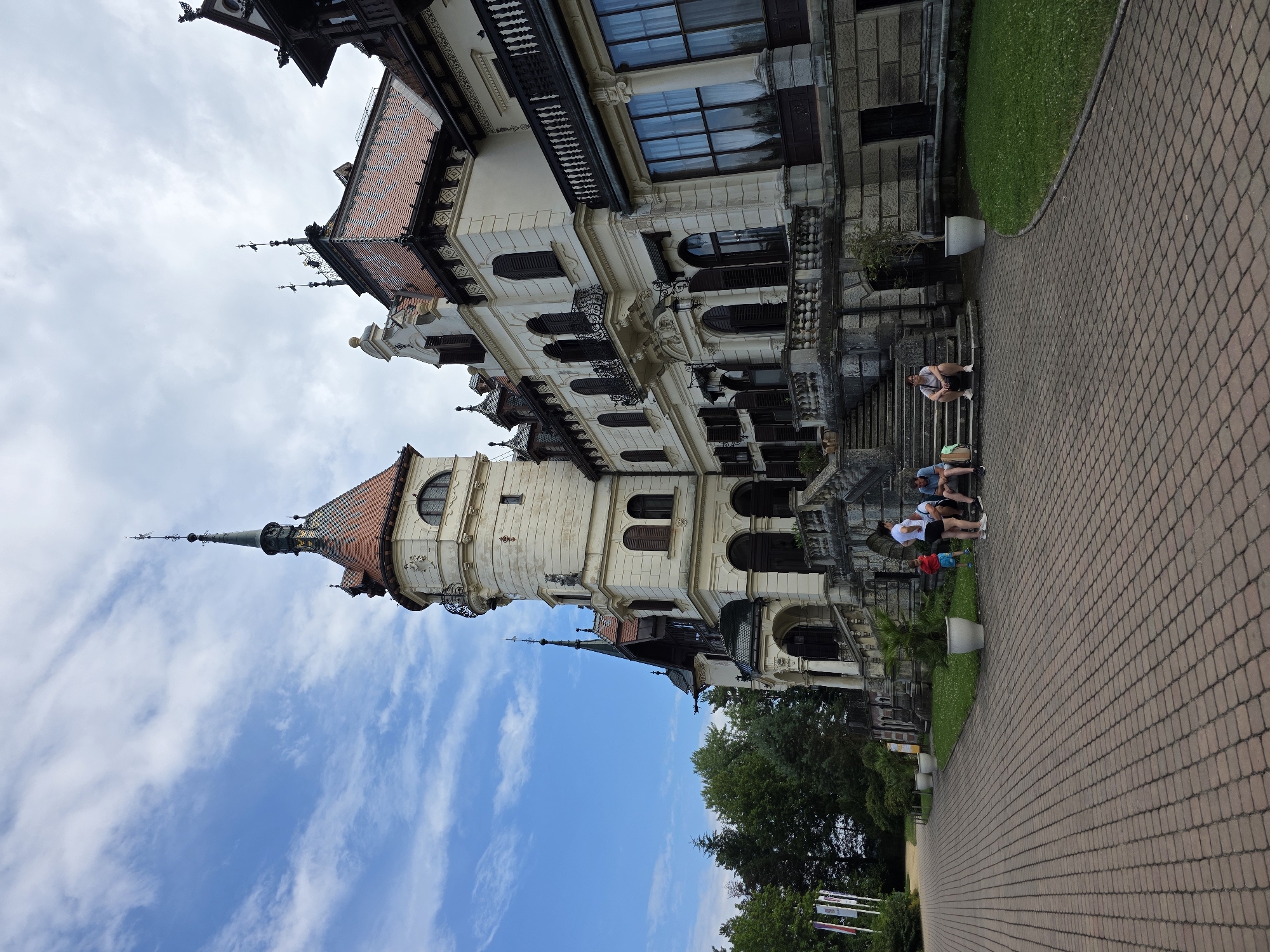 The image shows the exterior of a grand, ornate building, likely a castle or mansion, with a tower and multiple windows. The building is light-colored with red-tiled roofs and decorative elements. In front of the building, a paved walkway leads to a set of stone stairs where several people are seated. The sky is blue with scattered clouds. The building is surrounded by greenery, including trees and manicured grass. The scene appears to be a tourist attraction, possibly part of the **Lešná Zoo**, located near the city of Zlín, Czechia, as the architectural style resembles the chateau found on the zoo grounds.