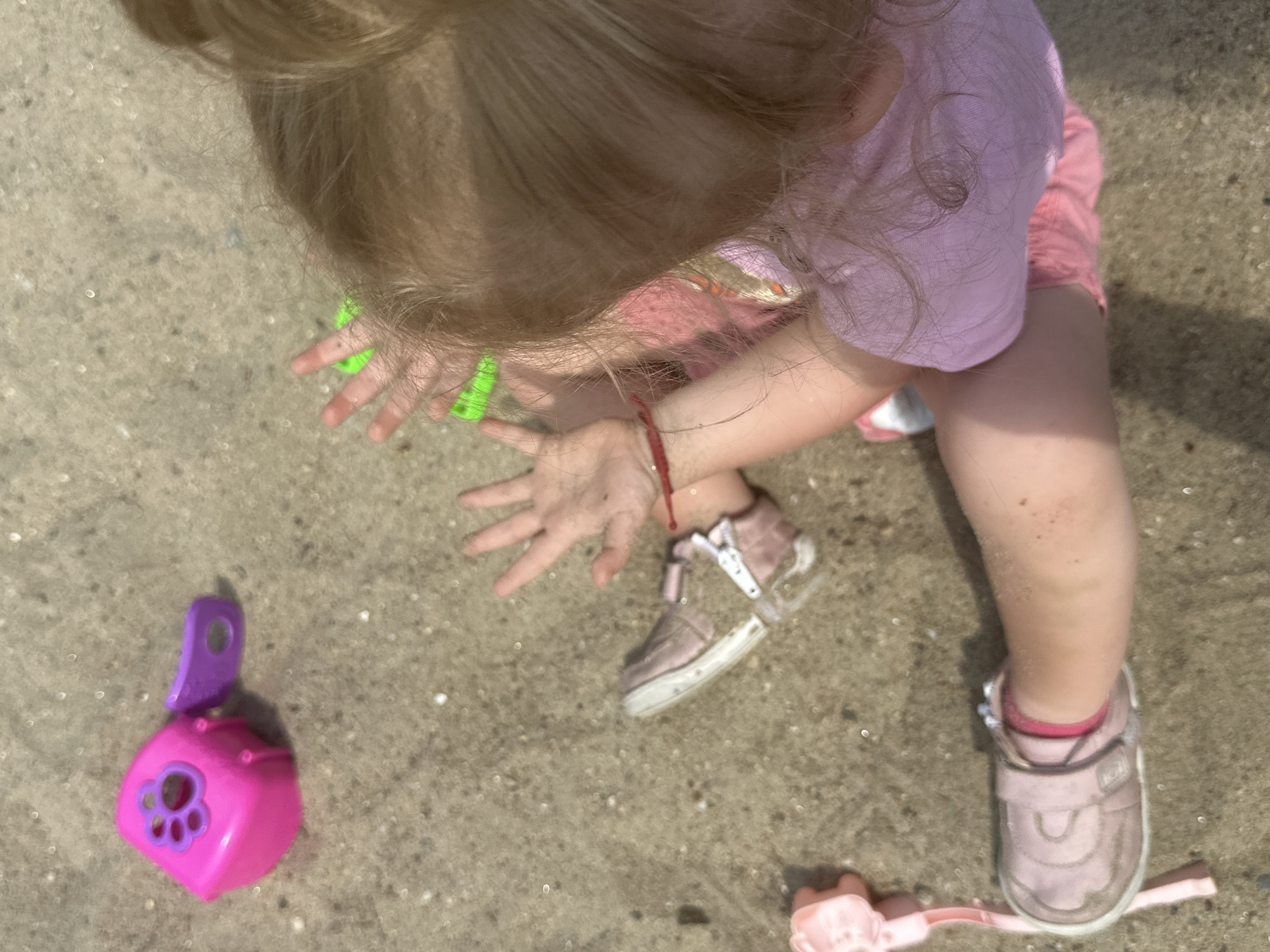A young child with blonde hair is sitting on a sandy surface. The child is wearing a pink t-shirt and pink shorts. One shoe, a pink sandal, is visible on their left foot, and the other is on the ground. Several toys are scattered around: a pink and purple plastic toy, a green plastic toy, and a pink plastic shovel. The scene appears to be outdoors, possibly in a park or playground, given the sand and toys. There are no other people or identifiable structures in the immediate vicinity. The lighting suggests it is daytime.