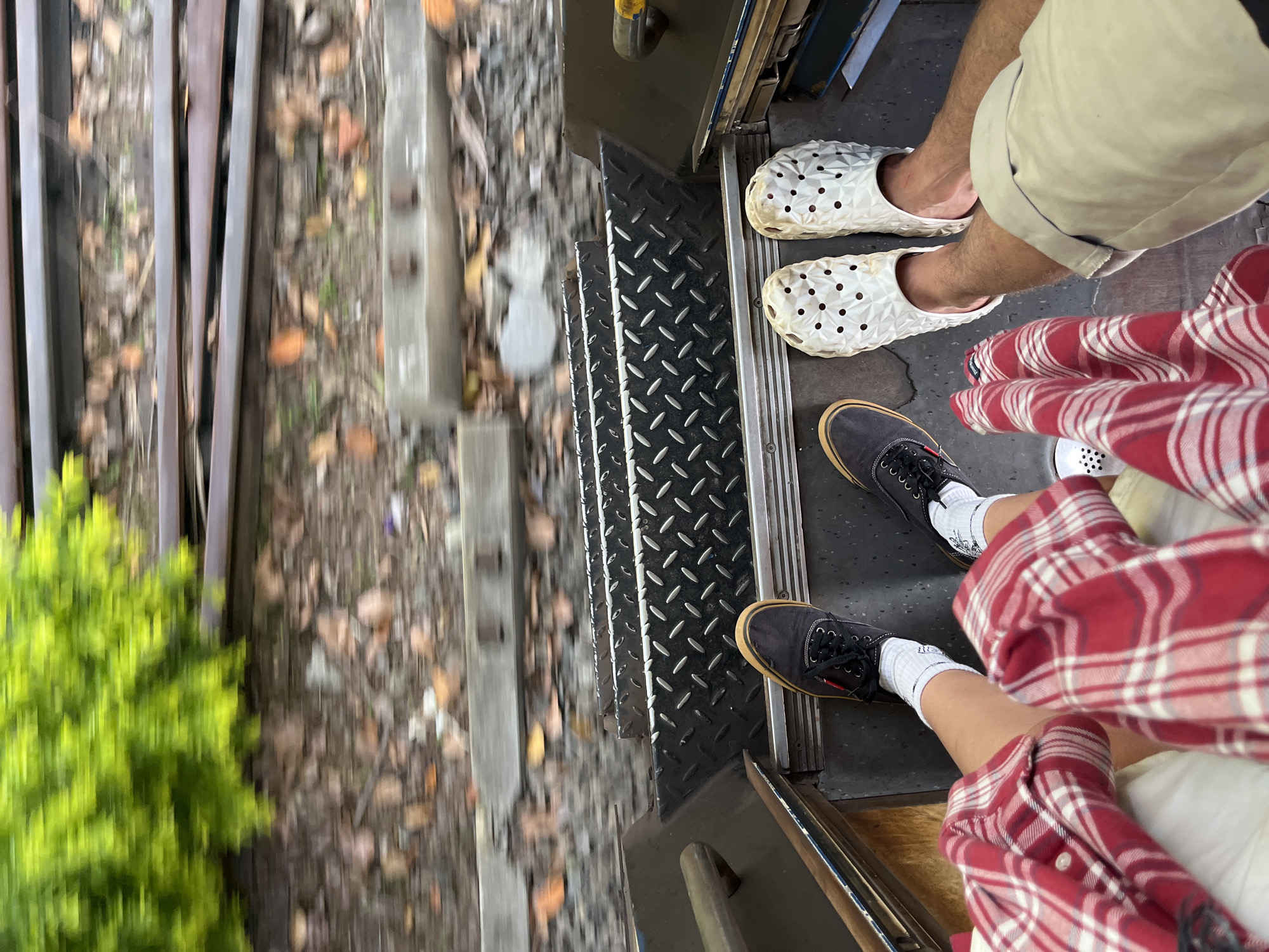 The image shows a top-down view of two pairs of feet standing on a metal staircase. The staircase is diamond-plated and appears to be on some form of transportation, possibly a train or tram, as suggested by the blurred railway tracks and foliage in the background. One person is wearing dark canvas shoes with white socks, and the other is wearing white Croc-like shoes. Both individuals are dressed in casual clothing, with one person wearing a red and white plaid shirt. The scene appears to be outdoors in a motion blur, indicating movement. Based on the provided context that the image is from Kalutara, Sri Lanka, it is likely that this scene is depicting a journey on a local train or possibly a tuk-tuk or other open-air vehicle, with the blurred background suggesting the lush greenery and tracks common in the region.