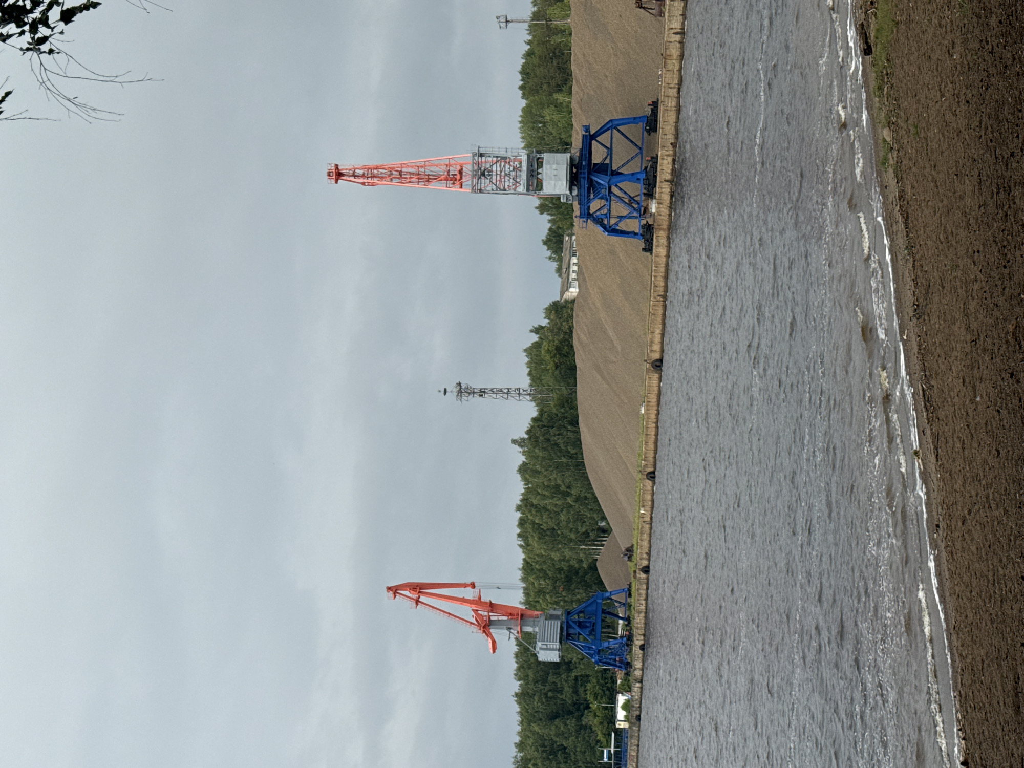 Two large industrial cranes, one orange and one orange and red, stand on a concrete pier next to a large pile of gravel. The cranes are positioned to the left and right of the frame, and the gravel pile stretches across the middle ground of the image. The water in the foreground is choppy and dark. A line of trees forms the background, with a cloudy sky overhead. A tall metal tower with a bird's nest structure on top is visible in the distance to the right of the frame. The scene appears to be a port or industrial area. This location is likely the Chaikovsky port on the Kama River, in Chaikovsky, Russia, known for its industrial activity and freight handling.