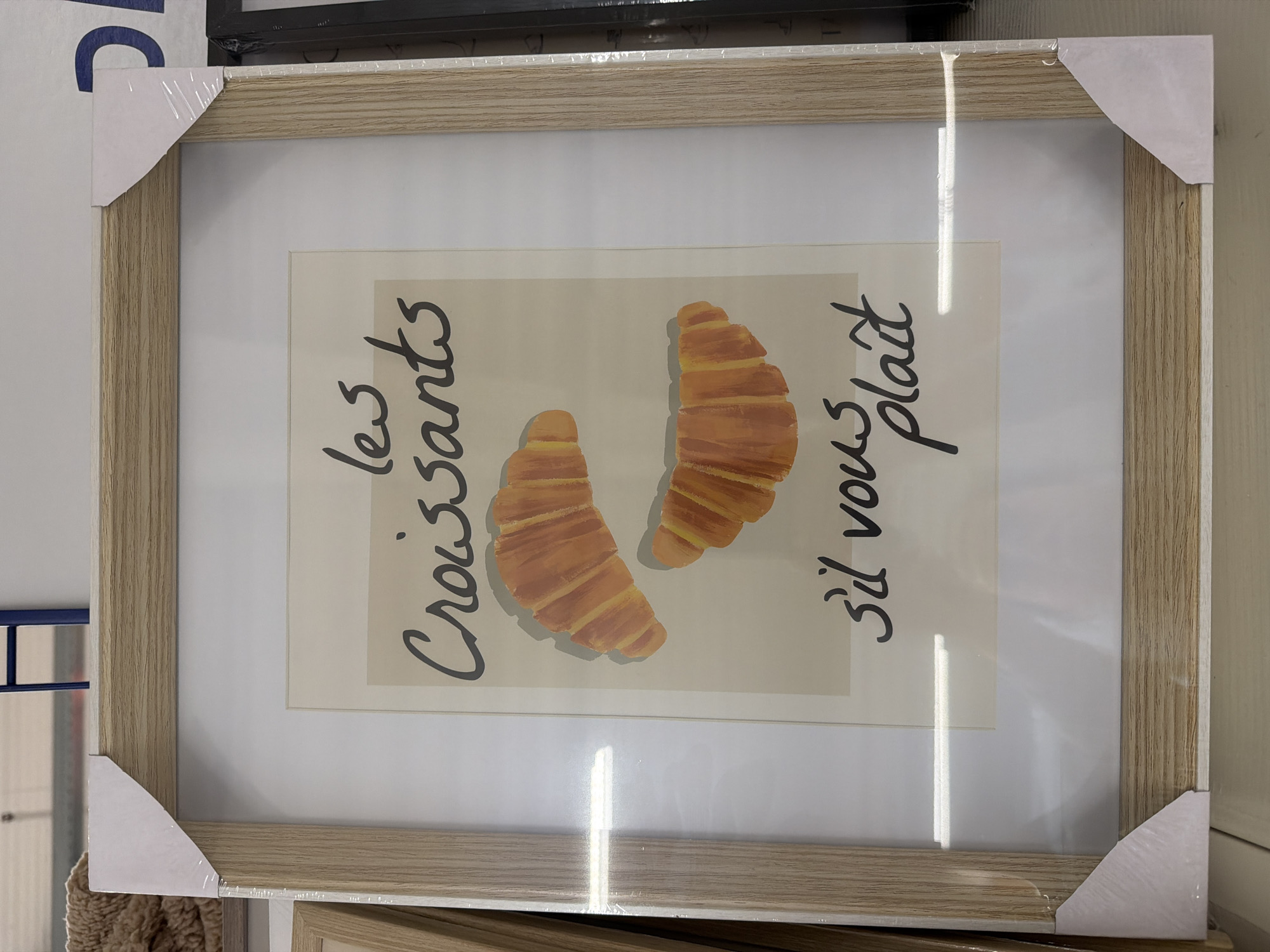 The image shows a framed print of two croissants. The print is inside a light-colored, wood-effect frame with protective corner pieces. The artwork depicts two croissants and the French phrase "Les croissants, s'il vous plaît" (The croissants, please). Based on the surrounding objects, the scene is likely located in a retail environment or store, possibly in the city of Luxembourg, Luxembourg. The frame and print appear to be a commercially available item for sale. Other items and store fixtures are partially visible in the background.