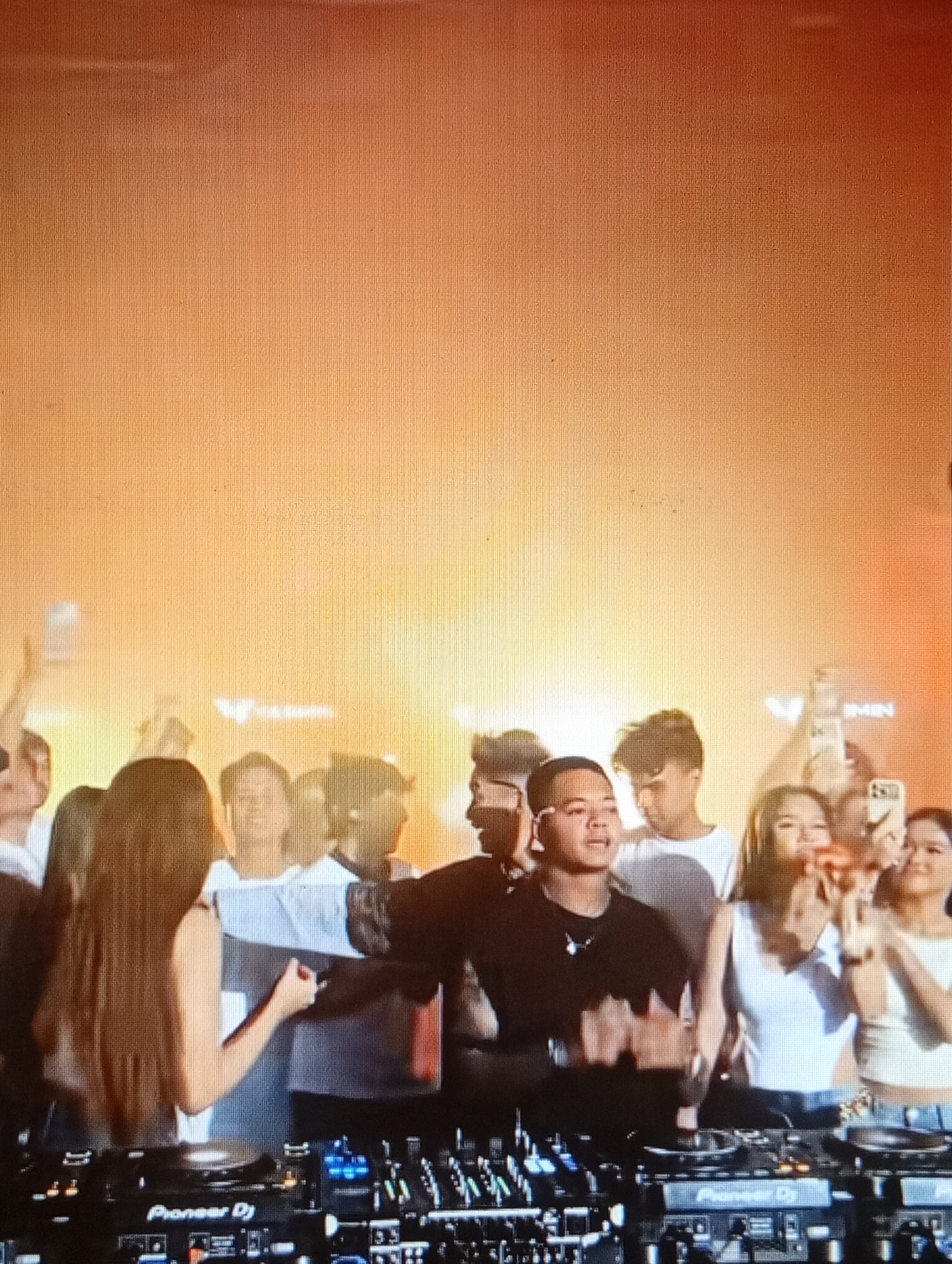 This media file shows a nighttime outdoor event, likely a concert or festival, with a DJ set in the foreground. The scene is illuminated by bright, warm orange and yellow stage lighting and fog, creating a vibrant and energetic atmosphere. A crowd of young people is visible behind the DJ, many of whom are holding up their phones, suggesting they are capturing the moment or interacting with the performance.
The central subject is a male DJ, wearing a black t-shirt, actively engaged with the DJ equipment, which is clearly labeled "Pioneer DJ". He appears to be controlling the music and interacting with the crowd. To his right, several women in the audience are also visible, some smiling and looking towards the stage. Their attire suggests a casual, festive event. The background is filled with the warm haze of stage lighting, making it difficult to discern specific details of the location. However, the presence of a crowd and a DJ setup strongly indicates a live performance environment. The overall mood is one of excitement and enjoyment, typical of a music event. There are logos visible on the backdrop which appear to read "WIMIN".