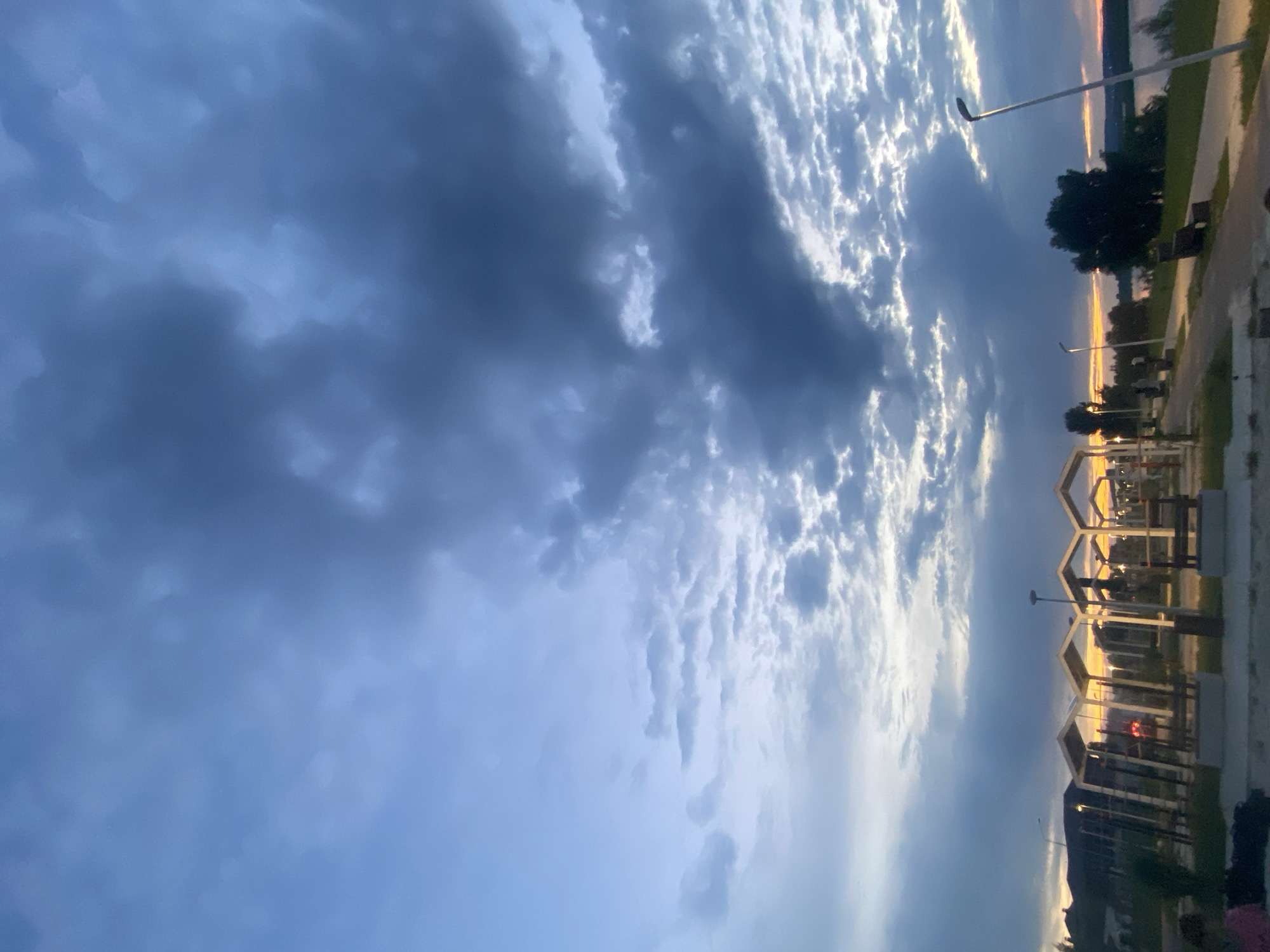 This is a vertical, outdoor, eye-level shot taken at dusk in Dyurtyuli, Russia. The sky is filled with a dramatic mixture of dark, ominous storm clouds and bright, fluffy white clouds illuminated by the setting sun. The light from the sun, which is low on the horizon, casts a warm, golden glow on the clouds and the distant landscape. The overall mood is one of anticipation, with the dramatic sky suggesting an approaching storm, but the warm light also conveying a sense of peace and beauty.
In the foreground, there are some modern, geometric structures that appear to be part of a park or public space. These structures are outlined by the setting sun, and the light creates a warm glow around them. There are also some streetlights visible, adding to the urban feel of the scene. In the background, a body of water is visible, likely a river or lake, with trees and the silhouette of a distant town or city on the opposite bank. The time of day is clearly dusk, as indicated by the low angle of the sun and the warm, golden light. The weather is mixed, with both dark storm clouds and bright, sunny clouds present. The location cues of Dyurtyuli, Russia, and the presence of a body of water suggest a potentially scenic or recreational area. There are no visible people or text in the image.