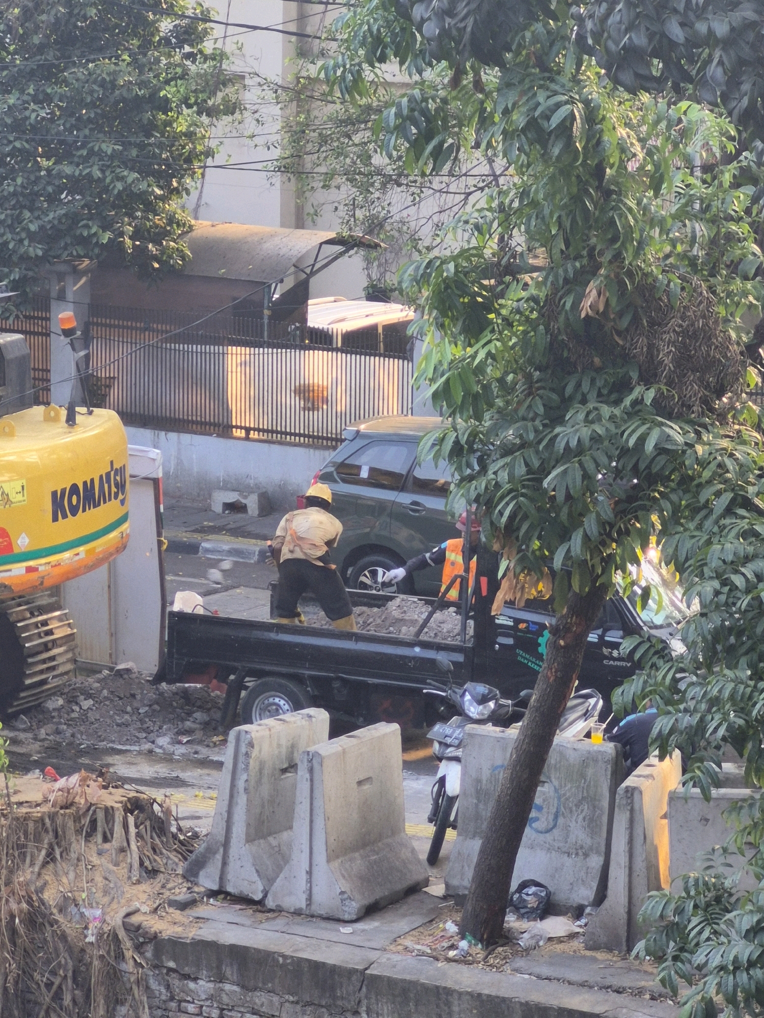 In Central Jakarta, Indonesia, a construction scene is depicted. A yellow Komatsu excavator is partially visible on the left. In the center, a construction worker wearing a yellow hard hat, a beige vest over a light-colored shirt, and dark shorts with yellow boots is standing in the back of a black pickup truck filled with gravel. He appears to be shoveling or moving the gravel. Another construction worker, wearing an orange safety vest, stands nearby. A dark green SUV is parked behind the pickup truck. A white motorcycle is parked to the right of the pickup truck. Several concrete barriers are placed along the roadside. Lush green trees with dense foliage dominate the foreground and frame the scene. The environment suggests an urban setting with ongoing infrastructure work.