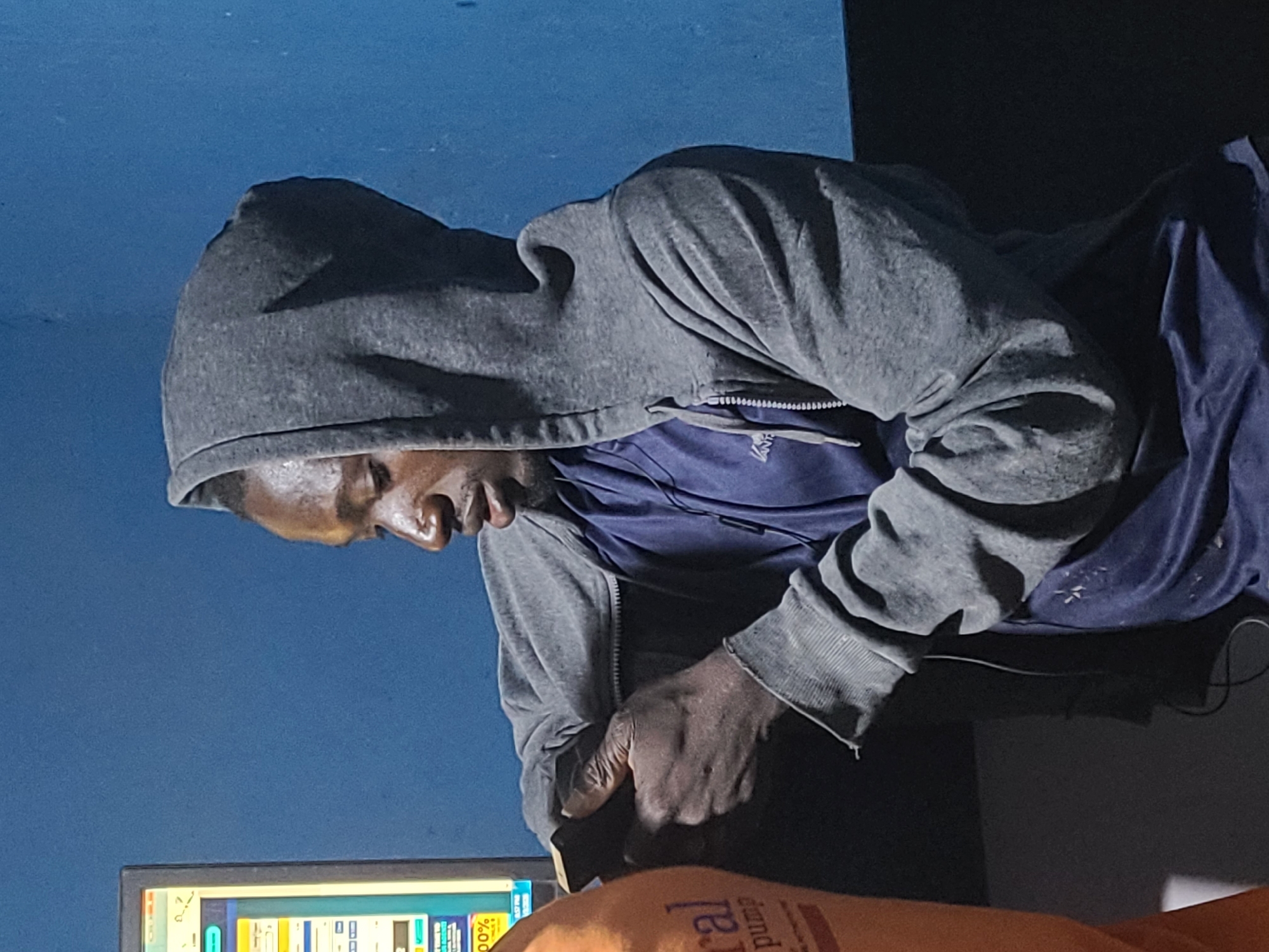 The sideways image features a person seated indoors, viewed from the side in profile, against a plain blue wall. The individual has dark skin and is wearing a grey hooded sweatshirt over a dark blue shirt. They are leaning forward, looking down with a focused or pensive expression.
Their hands are visible, appearing dark and somewhat rough, and they are holding or interacting with a dark, rectangular object, possibly a smartphone or a remote control. This object rests on a dark surface in front of them, which appears to be a desk or table.
On the desk, a computer monitor is visible, displaying a colorful graphical user interface with various elements, including what looks like columns, numbers, and possibly progress bars or indicators. Partially visible text on the screen includes "00%" and the number "07".
In the foreground, an orange object is partially visible, showing parts of text which read "ural pump," possibly from a product label such as "Natural pump." The grey hoodie worn by the person shows some texture and minor wear. The overall lighting suggests an indoor setting, possibly with low ambient light, illuminated partly by the computer screen.