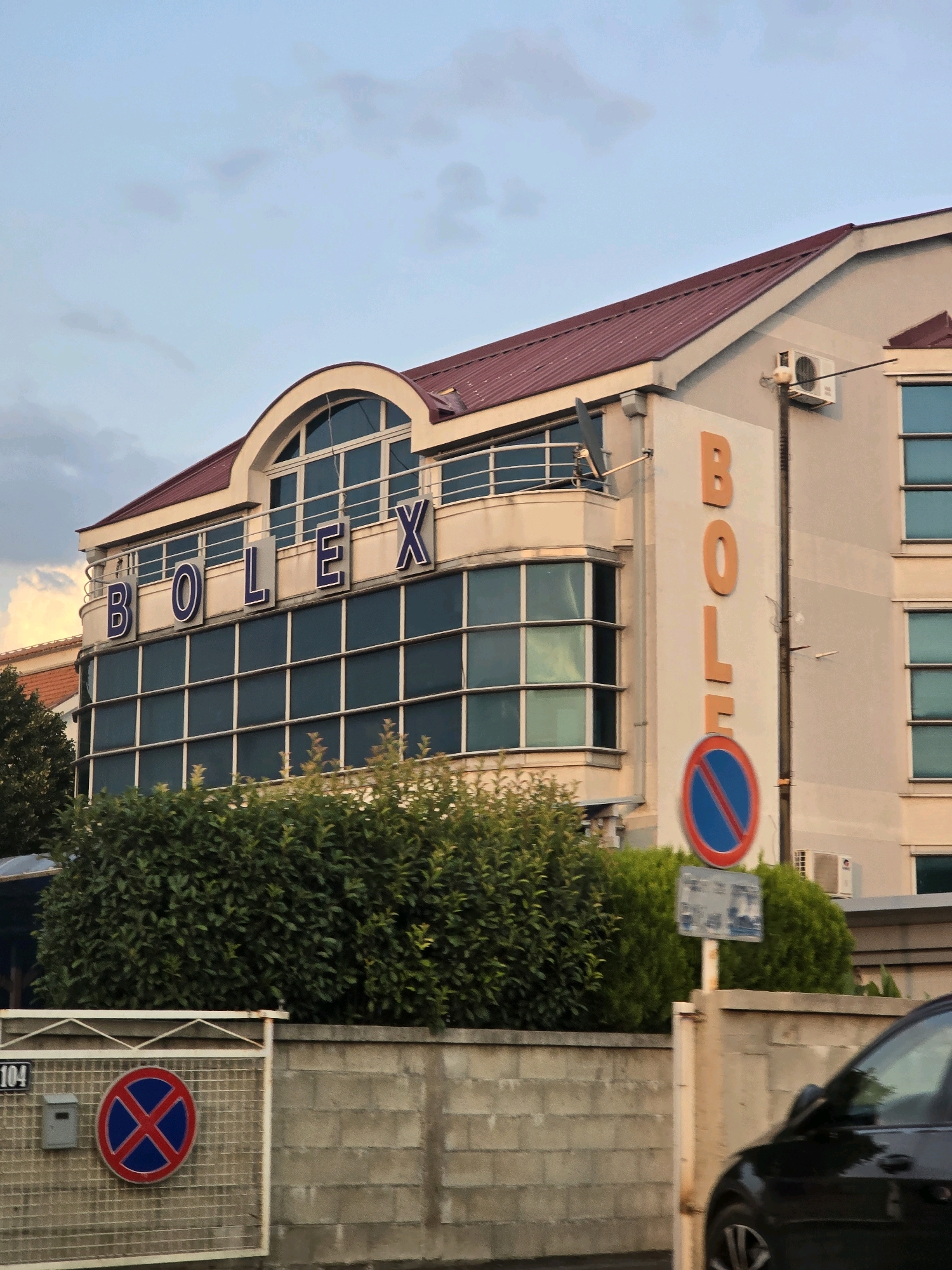 The image shows a building with the word "BOLEX" written on it in large blue letters. Below this, the word "BOLEX" is also visible in large orange letters vertically on the side of the building. A "no stopping" sign is affixed to the side of the building. In the foreground, a section of a gray stone wall with a metal fence is visible. A mailbox is attached to the fence, and a traffic sign with a red circle and blue background with a red 'X' inside is mounted on the fence. A black car is partially visible on the right side of the image. The building appears to be modern, with large windows. The sky is cloudy and appears to be either early morning or late evening. This is located in Podgorica, Montenegro.