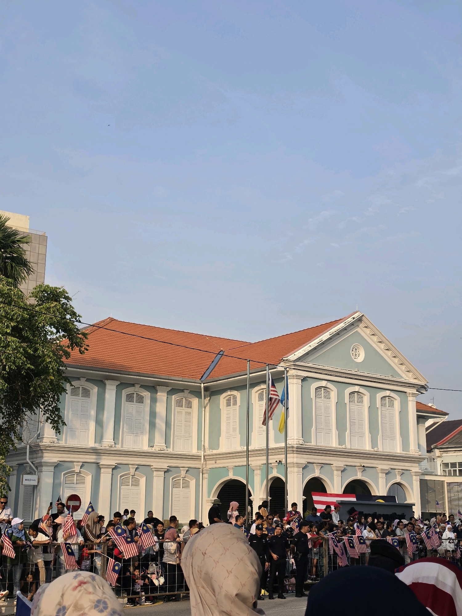 In George Town, Malaysia, a crowd gathers in front of a light blue colonial-style building with white shutters and an orange tiled roof. The building features arched doorways and decorative white trim. Several Malaysian flags are visible, held by people in the crowd and also flown from poles in front of the building. Some individuals are wearing headscarves. The sky is clear and light blue. The scene appears to be a public gathering or event.